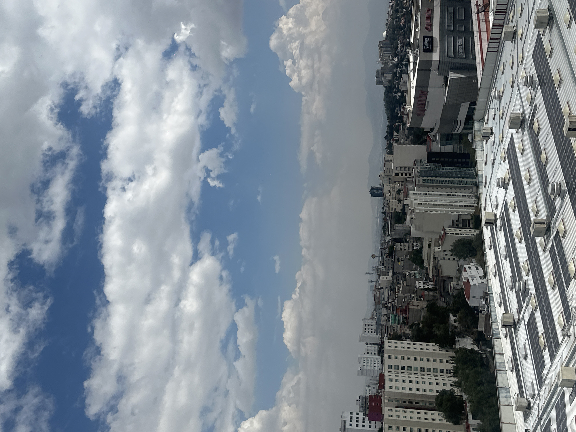 The image depicts an aerial view of Huixquilucan de Degollado, Mexico, with the sky occupying the left half of the frame filled with cumulus clouds and blue sky. The city sprawls across the right half. In the foreground, a metal roof structure dominates, equipped with rows of air conditioning units and what appear to be solar panels. Further into the city are densely packed buildings, primarily white and gray, interspersed with green spaces. Some taller commercial buildings can be distinguished, including one with visible signage. In the background, mountains are visible through a haze or smog.