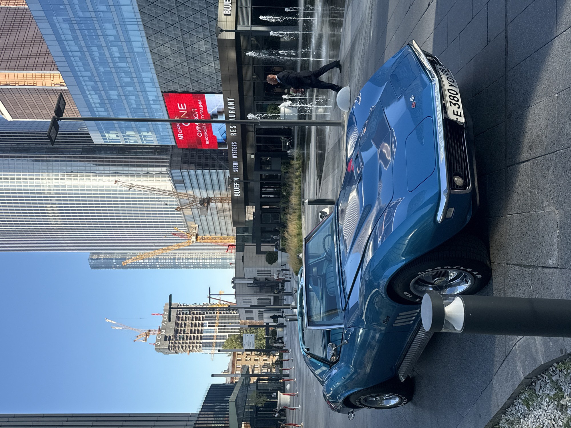 A bright blue 1969 Chevrolet Corvette Stingray convertible is parked on a paved surface in front of a modern building complex. The car is angled towards the right side of the frame, with its front end prominent. In the background, several skyscrapers of varying architectural styles, some under construction with visible cranes, dominate the skyline. A large digital billboard displays red and white text. To the right, a man in a dark suit walks past a fountain with several water jets. The environment appears to be an urban area in the city of Khimki, Russia, possibly a commercial or business district, with a clear blue sky overhead.