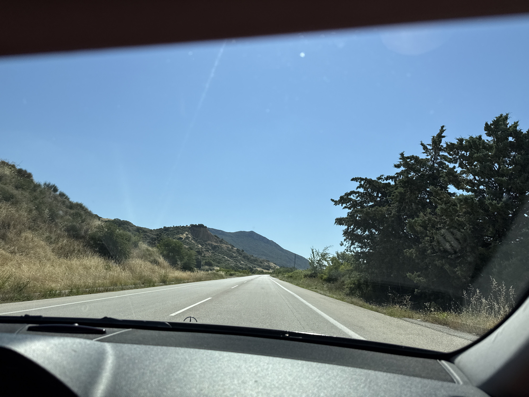 The image is a first-person perspective shot from inside a vehicle, looking out through the windshield at a road leading into a landscape. The road is paved and appears to be a two-lane highway with a solid white line marking the right edge and a broken white line to the left. The sky is clear and blue.
On the left side of the road, there is a gently sloping hillside covered in dry, golden-brown grass and scattered green shrubs. Further back on the left, the terrain becomes more rugged with rocky outcrops and sparse vegetation.
On the right side of the road, there are dense evergreen trees, possibly cypresses or junipers, growing close to the roadside. Beyond the trees, the landscape rises into rolling hills and mountains covered in greenery.
The foreground of the image shows the dashboard of the vehicle, with parts of the steering wheel and windshield wipers visible. The lighting suggests it is a sunny day, likely midday or early afternoon. The overall scene depicts a scenic drive through a rural, mountainous area.