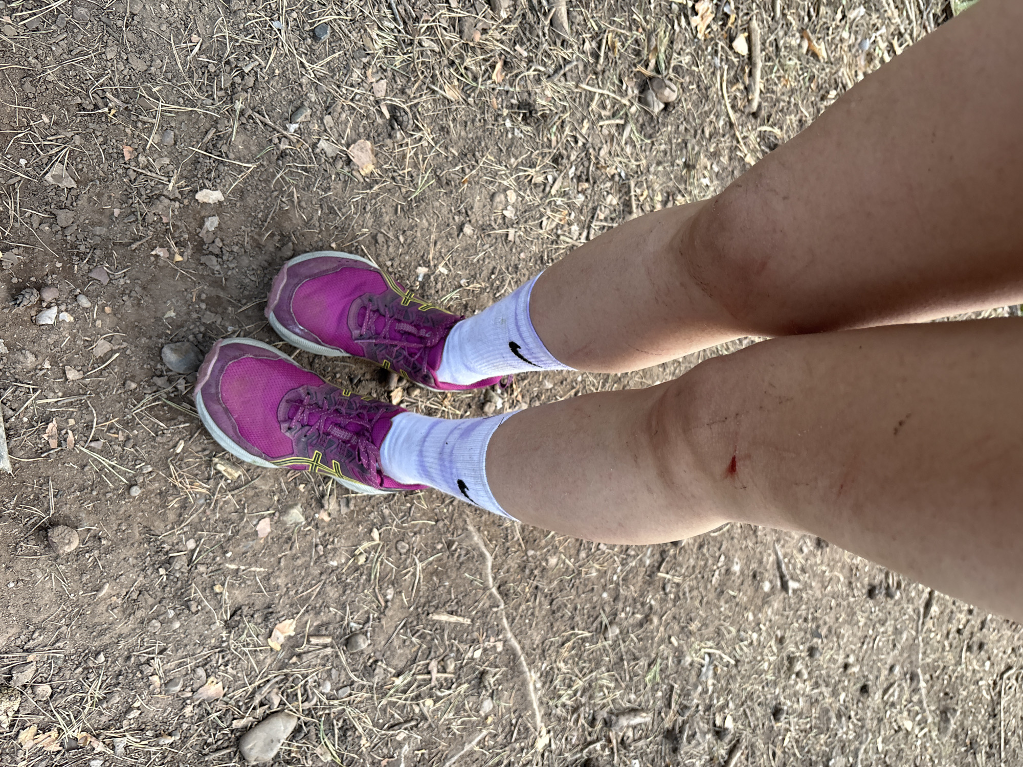 A first-person overhead view shows a person's legs and feet on a dirt ground. The person is wearing pink running shoes with yellow accents and white socks with a black Nike swoosh logo. The ground consists of dirt, small rocks, and dry pine needles and twigs. There are minor abrasions visible on the person's right leg. There is no discernible indication of the location being Perm, Russia.