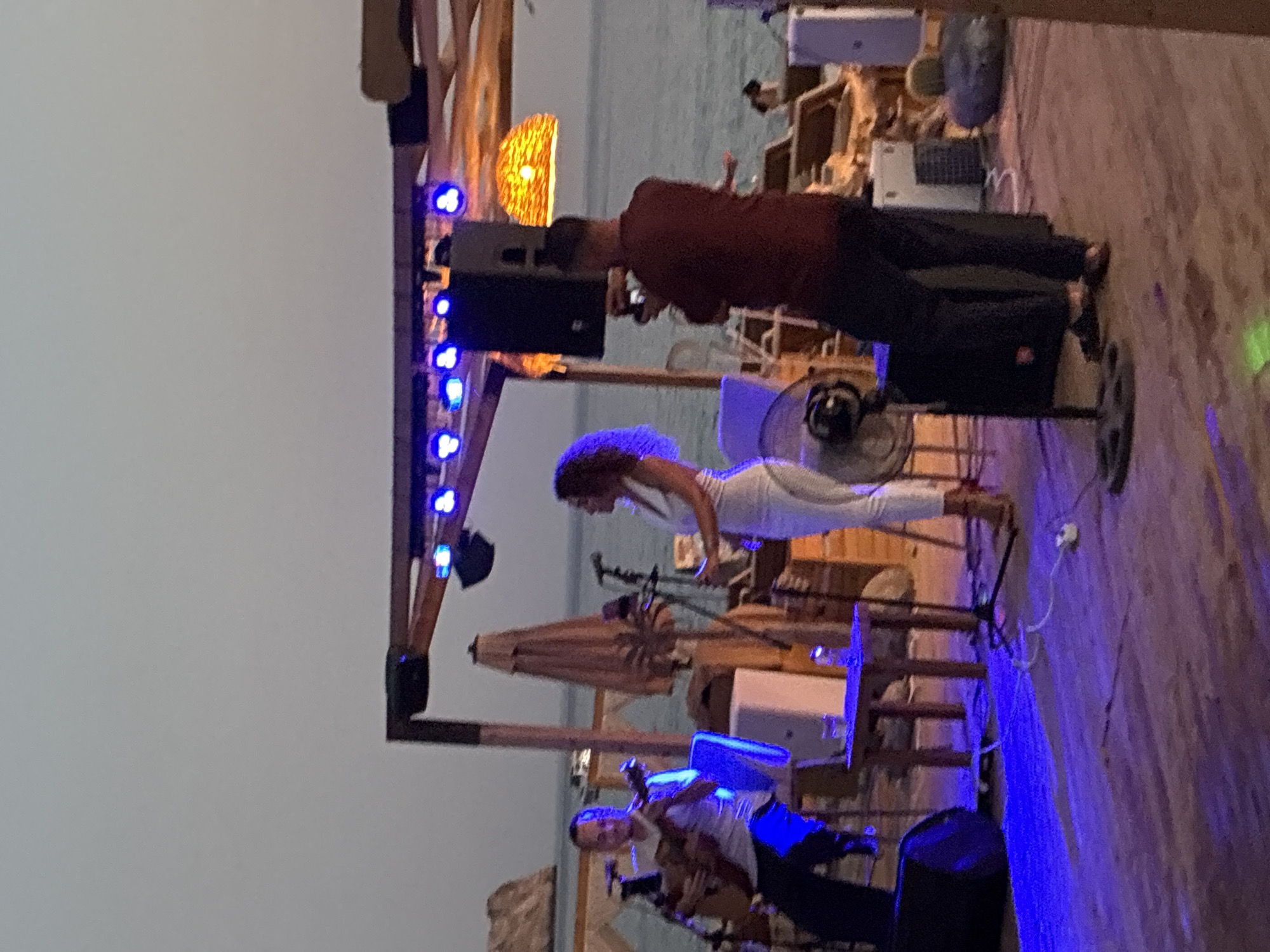 An outdoor evening performance is taking place on a wooden deck by the sea, likely in Alanya, Turkey. A male musician is seated on the left, playing an acoustic guitar. A female singer, dressed in white, stands center-stage, facing right. A man in a maroon shirt is positioned behind the singer, appearing to film the performance with a camera or phone. The scene is illuminated by blue stage lights and a warm, woven light fixture hanging overhead. Speakers and a fan are visible on the stage. In the background, lounge chairs and a beach are discernible, with a rock formation in the distance. The overall atmosphere suggests a relaxed, open-air concert or event.