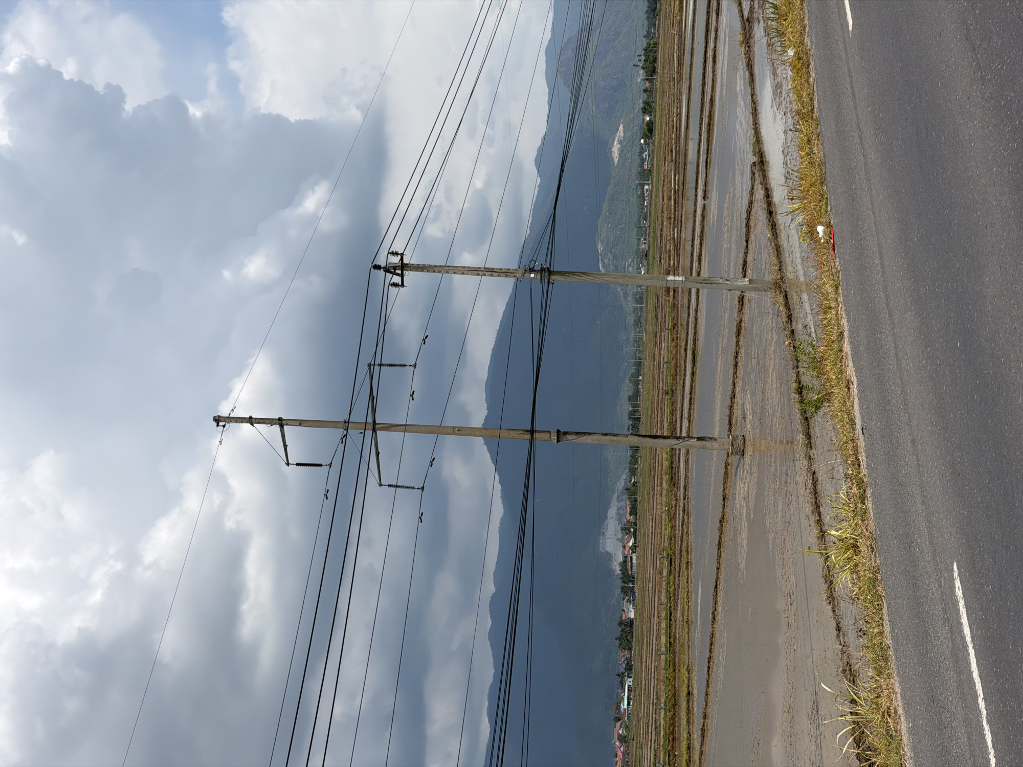 In Dien Lac, Vietnam, a rural landscape is depicted under a cloudy sky. The foreground features a paved road with a white line marking its edge, bordered by a strip of dry grass and some scattered weeds. Beyond the road lies a flooded field, divided into rectangular sections by raised earthen berms. Several utility poles with multiple wires stretch across the scene, supporting the overhead electrical infrastructure. In the middle ground, a small village with houses and buildings is visible, nestled against a backdrop of lush, green mountains. The overall impression is one of a tranquil, agrarian setting with the presence of modern infrastructure.