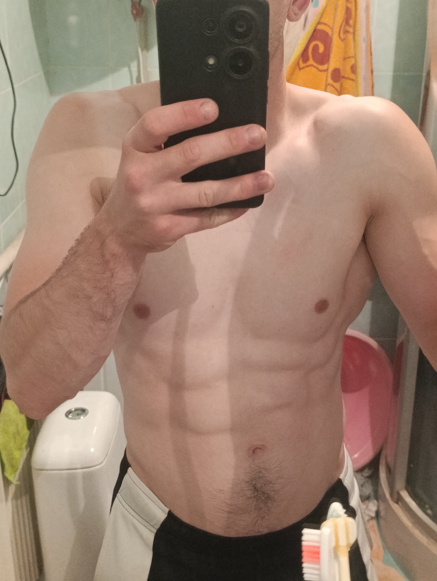 The image shows a close-up view of a person's torso, from the shoulders down to the upper thighs. The person is in what appears to be a bathroom, with tiled walls and a toilet visible in the background to the left. A black smartphone is being held in front of the person's chest, obscuring their face. The person's abdomen displays well-defined abdominal muscles. To the right, a pink basin or bucket is partially visible, along with a toothbrush in the foreground. The overall lighting suggests an indoor setting. There is no indication of the city of Fastiv, Ukraine.