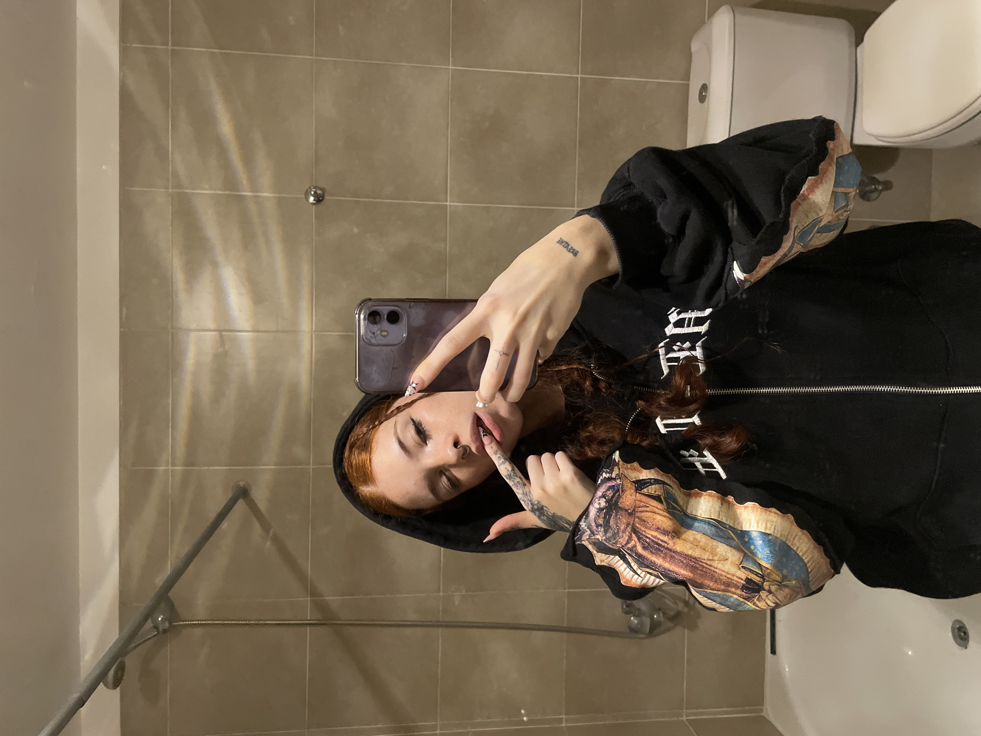 The image, rotated 90 degrees clockwise, shows a person taking a mirror selfie in what appears to be a bathroom, likely in Buenos Aires, Argentina.
The subject is a young individual with reddish-brown, wavy hair, wearing a black hooded sweatshirt. The sweatshirt features intricate, multi-colored artwork on its sleeves, depicting figures resembling the Virgin Mary in a classical art style, along with stylized, unreadable white text on the body. The person has a nose piercing, long fingernails with detailed designs, and a tattoo on their right wrist (appearing on the left in the mirror image) that reads "BEYVLIE" or "BEYTLIE" in a stylized font. They are holding a purple-cased smartphone to capture the photo, posing with their fingers near their mouth and eyes partially closed.
The background consists of light-colored, square-tiled walls with visible grout lines. To the right, a white toilet and a wall-mounted white fixture (possibly a toilet paper dispenser) are partially visible. Metallic fixtures, such as a shower rod or towel bar, are also present, reflecting the indoor artificial lighting. These light sources create distinct, diagonal rainbow-like refractions and patterns on the tiled wall. The scene indicates an indoor setting, consistent with a domestic or public restroom.