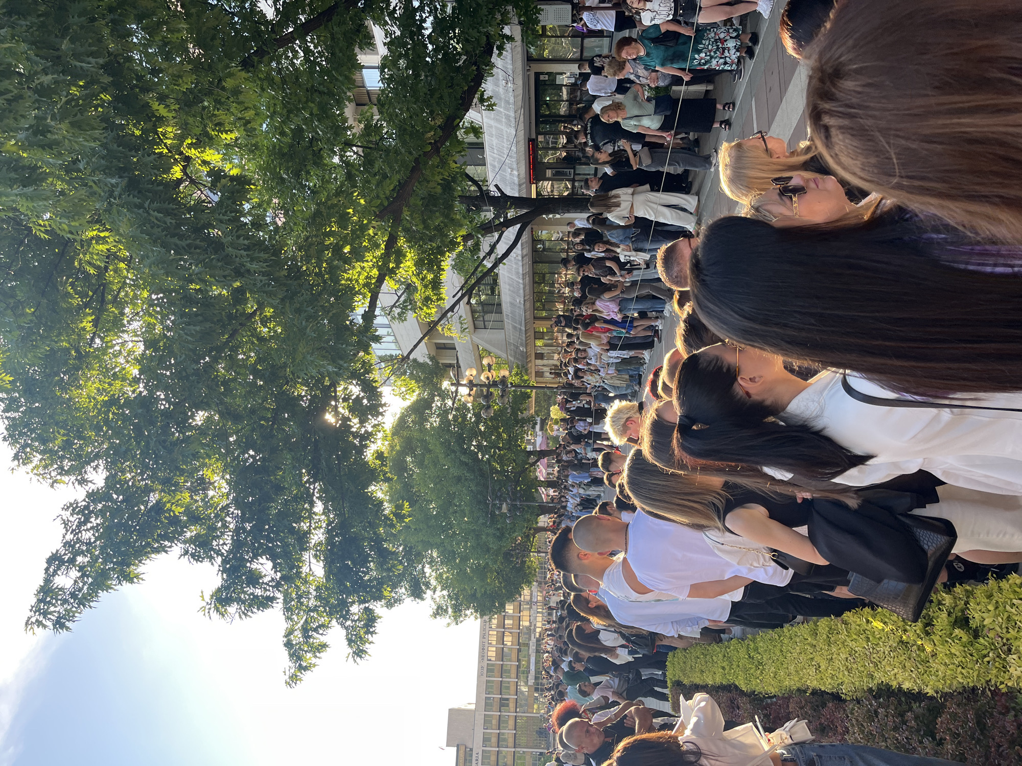 A daytime outdoor scene shows a dense crowd of people lined up along a city street in Blagoevgrad, Bulgaria. The view is partially obscured by the backs of individuals in the foreground. The crowd stretches into the distance with buildings and trees lining the street. Sunlight filters through the trees. People in the crowd are wearing a variety of clothing, including white shirts and casual attire. A low hedge or planting bed is visible in the lower right corner of the frame.