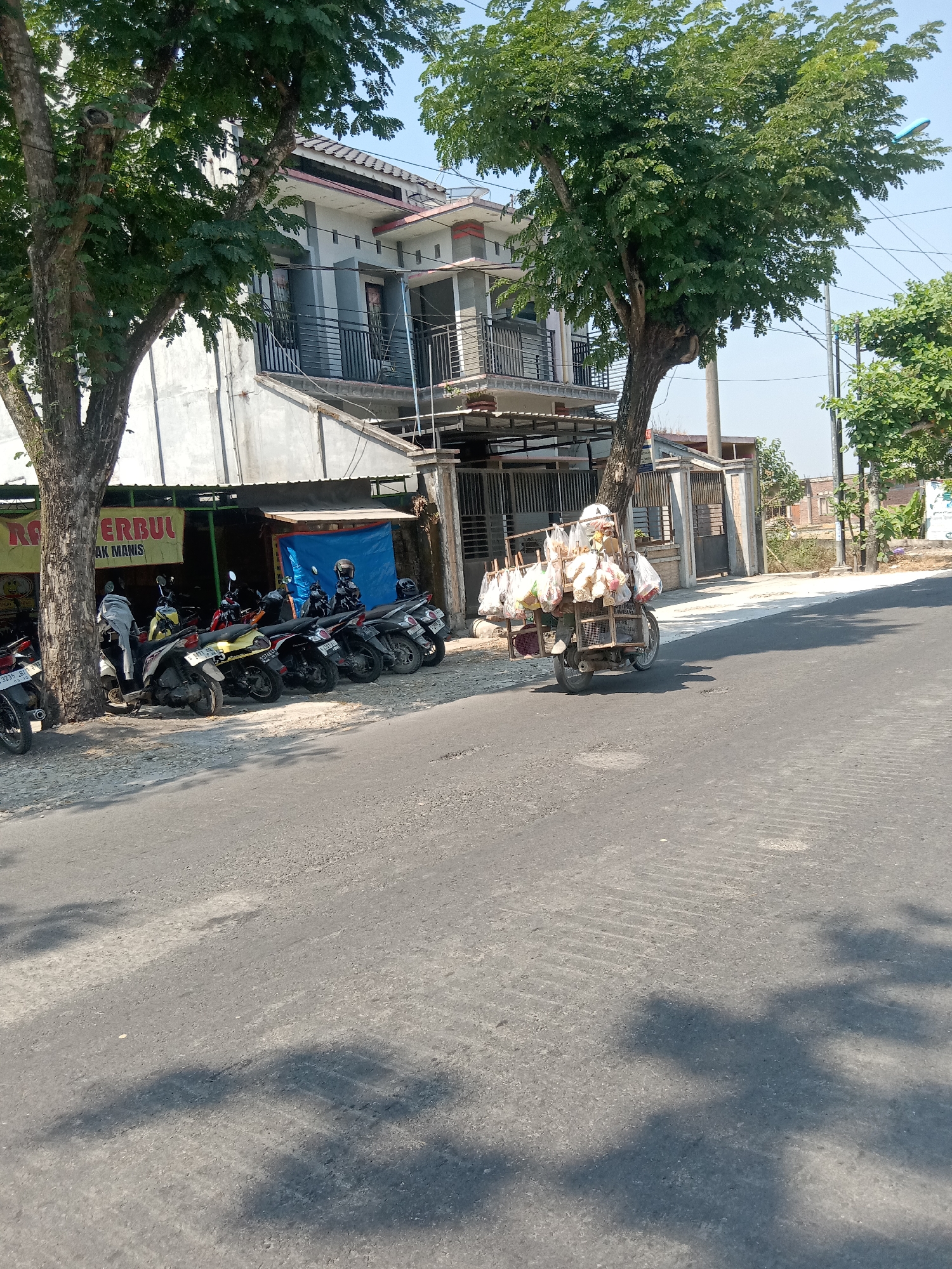 A street scene in Kemamang, Indonesia, features a row of parked motorcycles and a street vendor on a motorcycle cart. The vendor, wearing a helmet, is pushing a cart laden with numerous plastic-wrapped items, likely goods for sale, down the asphalt road. The cart is attached to a motorcycle, and the vendor is in the process of moving, indicated by the slight blur of the vehicle and the shadow it casts.
To the left of the road, a building with a gray facade and balconies is visible, partially obscured by a large tree. In front of the building, more motorcycles are parked, suggesting a commercial area or residential complex. A yellow banner with red text is also present, further indicating a business. The environment is sunny, with clear blue skies and shadows cast by the trees onto the road, indicating a bright day. The road surface appears somewhat worn, with visible texture.