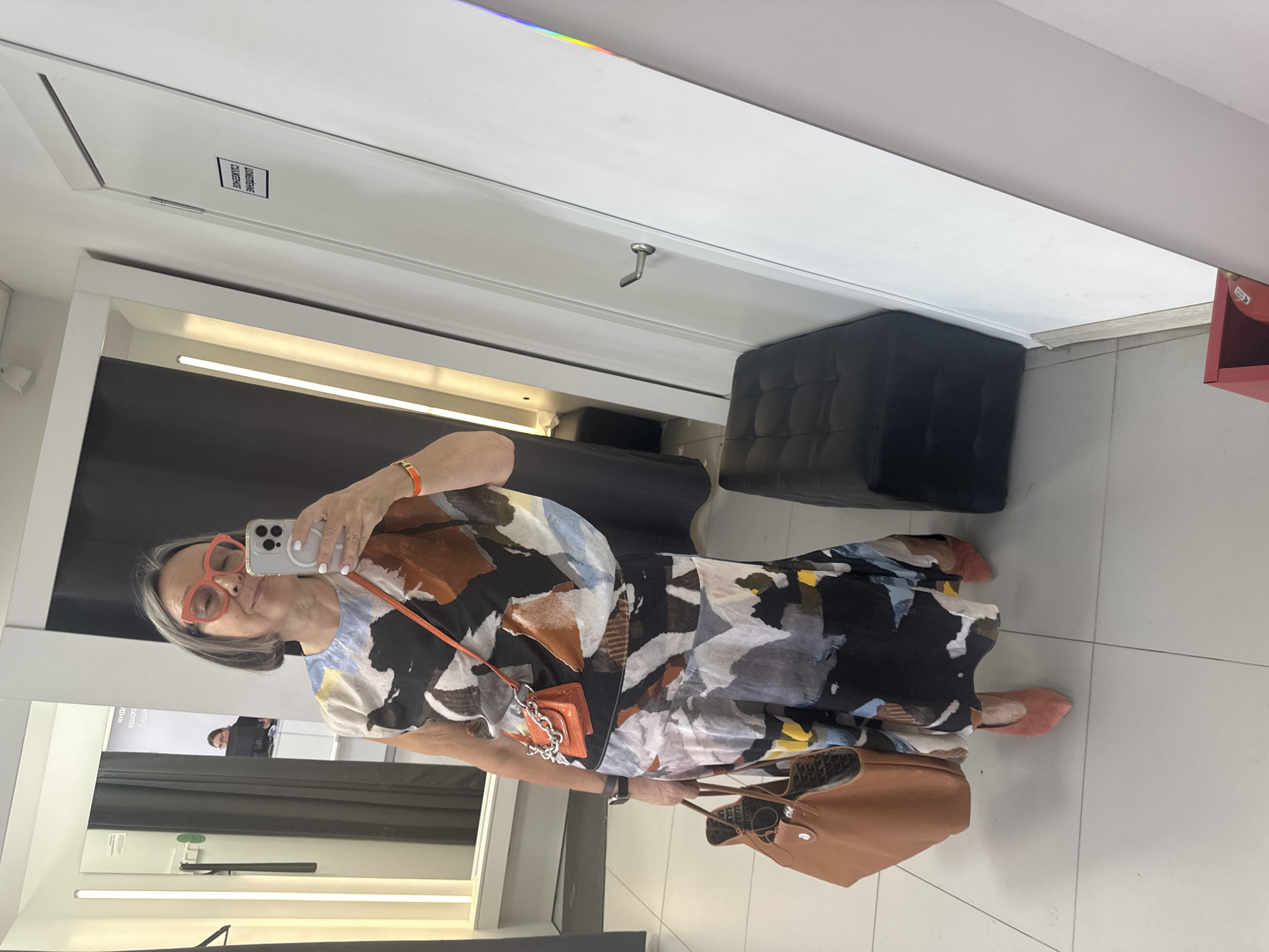 In a fitting room, a woman with gray hair, wearing oversized red glasses, a patterned dress, a small orange handbag, and suede shoes, takes a selfie with her phone. She holds a large tan tote bag in her left hand. The room has bright lighting, a large mirror, and a black ottoman. A sign with text in Cyrillic is visible on a white door. Another person is reflected in the mirror in the background, seated at a desk.