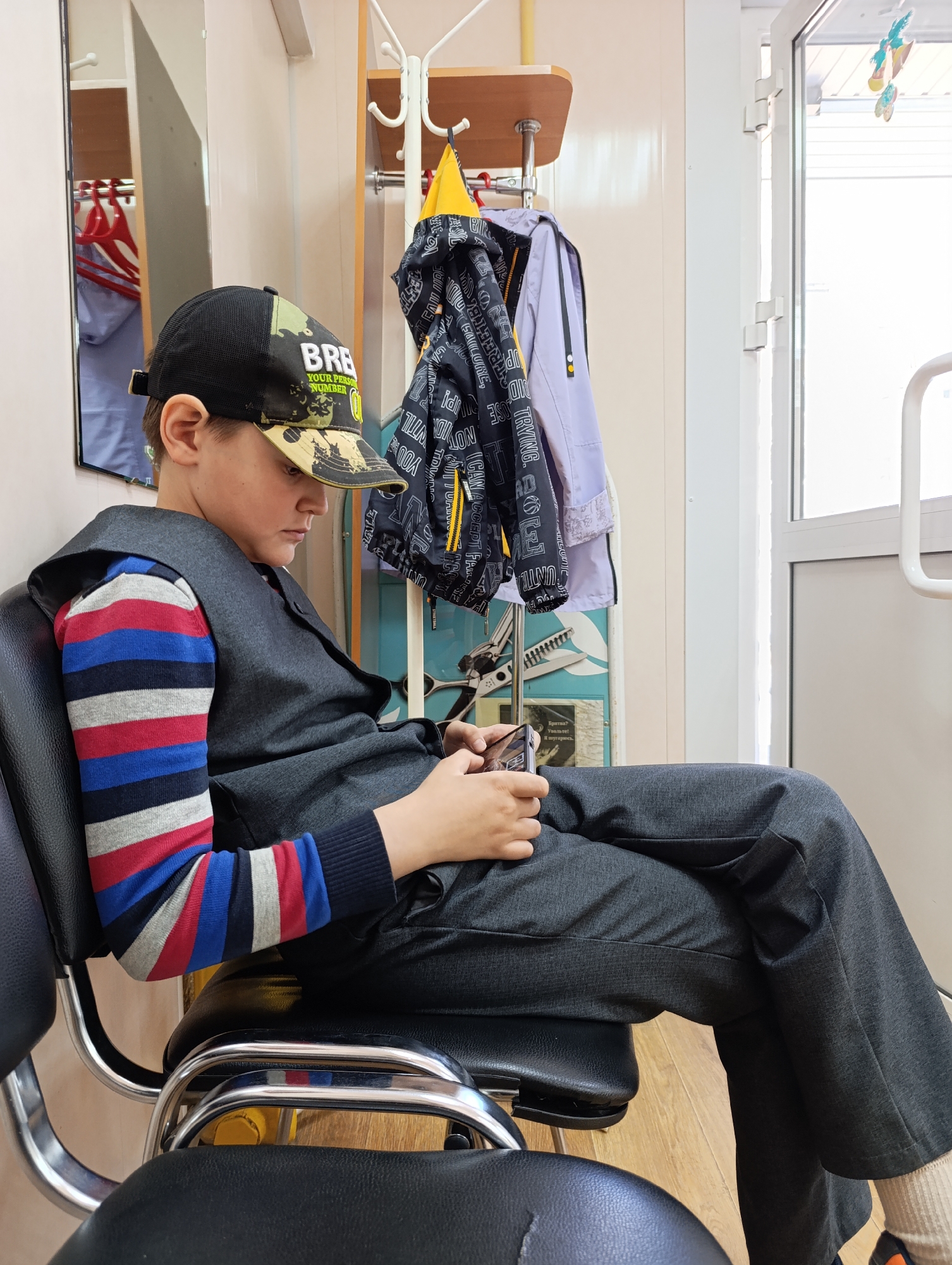 A boy sits in a black chair, focused on a mobile phone in his hands. He wears a black and camouflage-patterned baseball cap, a striped long-sleeved shirt with red, white, and blue stripes, and a dark gray vest. His legs, clad in matching dark gray pants, are crossed. A coat rack stands behind him, holding several pieces of outerwear. The environment appears to be indoors, near a white door with a window and a mirror reflecting clothing. Part of a Russian language message can be seen on the wall. The image was likely taken in Chaikovsky, Russia.