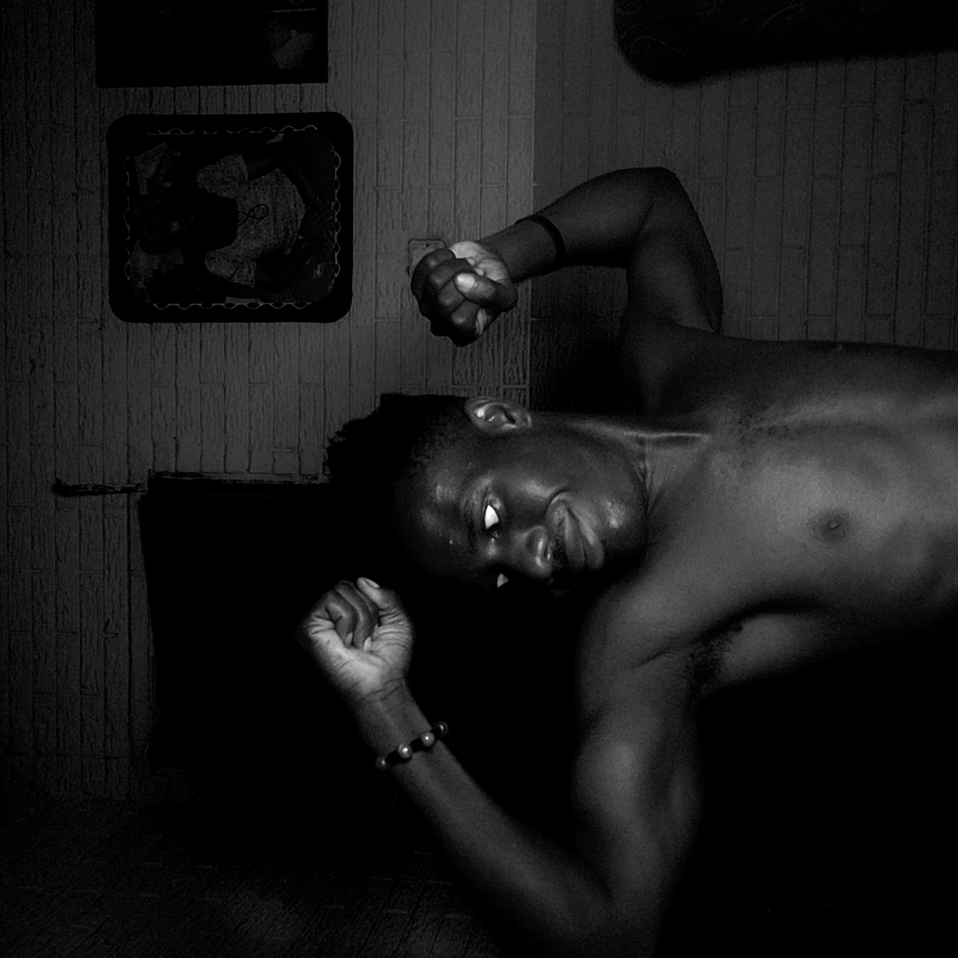 A black and white photograph depicts a shirtless Black man lying on his side in a room. His head is tilted towards the viewer, and his eyes are visible. One fist is raised above his head, while the other rests near his body, adorned with a beaded bracelet. Sweat glistens on his face and chest. The room's background includes a textured wall with two framed pictures hanging on it, and a dark, indeterminate object that partially blocks the view. The location is Narayi, Nigeria.