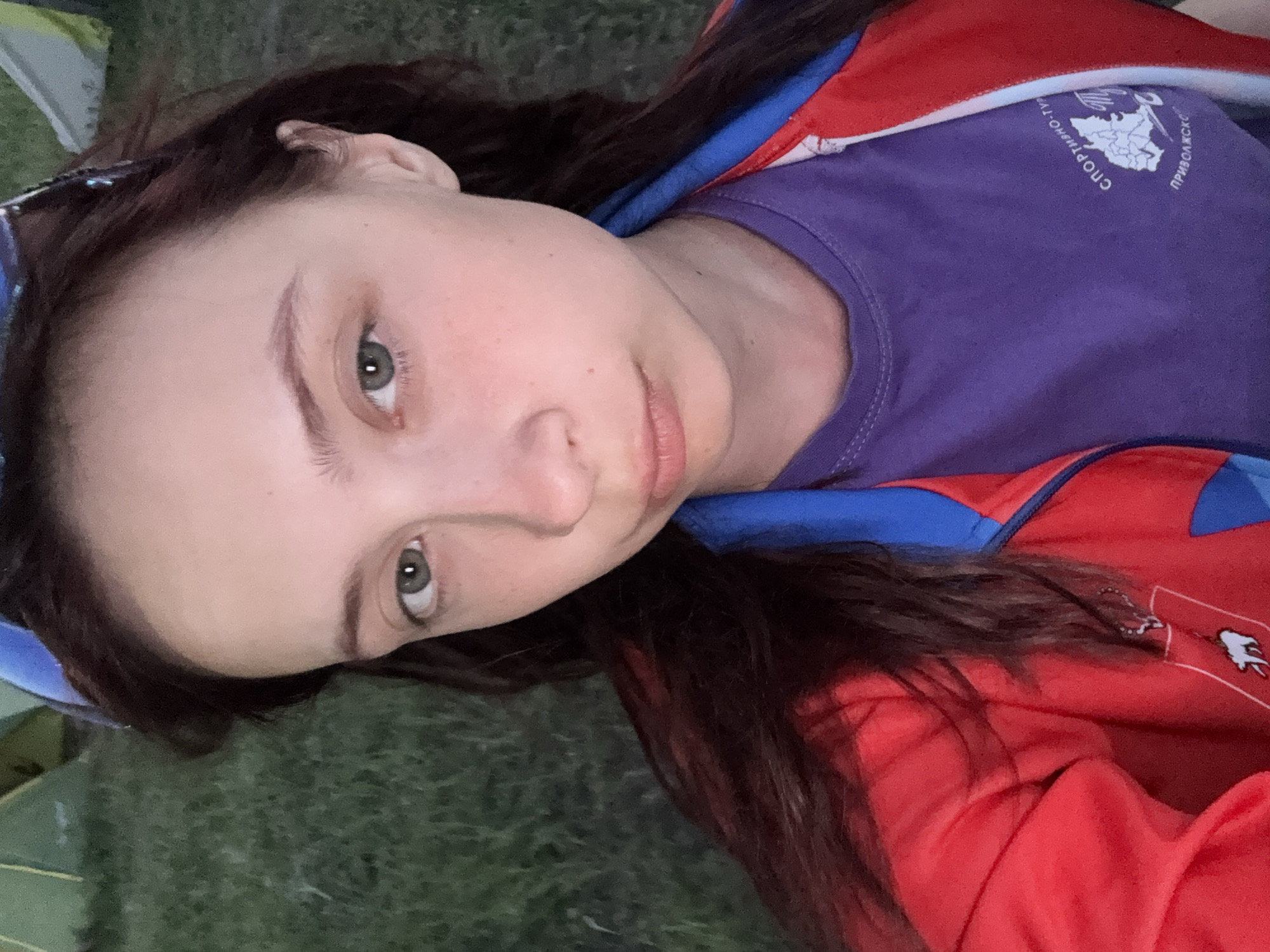 A young woman with brown hair and fair skin poses for a photo outdoors. She is wearing a purple t-shirt with the Russian words "Спортивно-трудовой ПРИВОЛЖСК" printed on it. A red jacket with blue trim is also visible. The background appears to be grass. The location is potentially Khvalynsk, Russia, given the text on the t-shirt.
