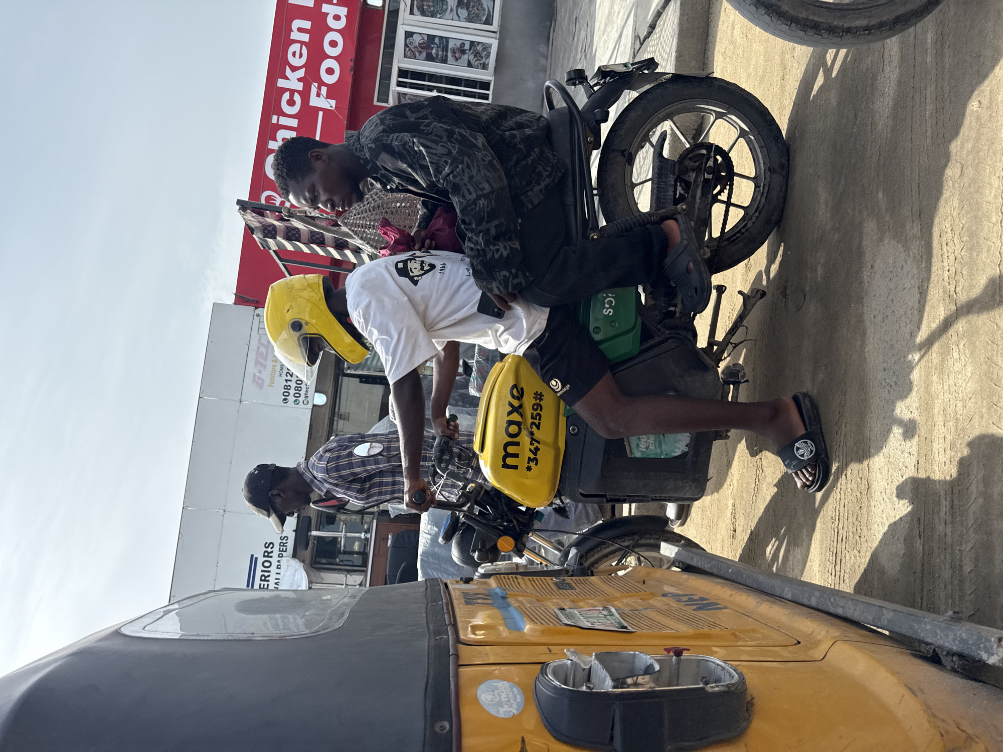 The image shows two young men on a motorcycle in Ado, Nigeria, near a "Chicken Food" restaurant. The driver wears a yellow helmet and white T-shirt with a logo, and the passenger wears a patterned shirt. The motorcycle has a yellow "maxe" branded delivery box. A yellow tricycle, partially visible, is in the foreground. Storefronts advertising furniture and interior decorations are in the background.