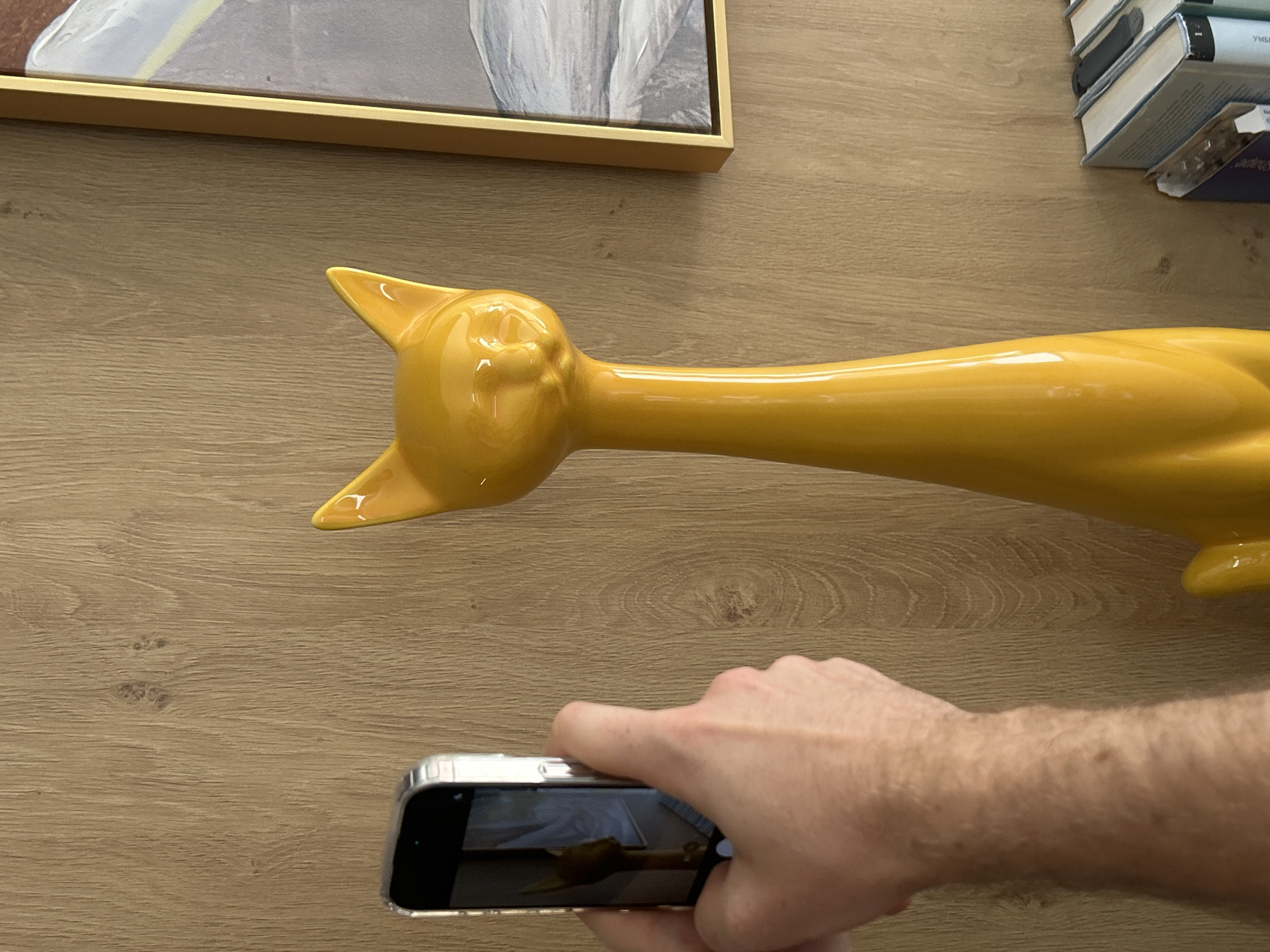 A person is holding a smartphone in their right hand, taking a picture of a yellow ceramic cat figure. The cat figure is lying on a wooden surface, oriented horizontally, with its head toward the upper left corner of the frame and its tail extending toward the lower right corner. Above the cat's head is a framed picture with a gold frame. In the upper right corner, there is a stack of books. The wooden surface appears to be a tabletop or a desk. The location cannot be determined from the image.