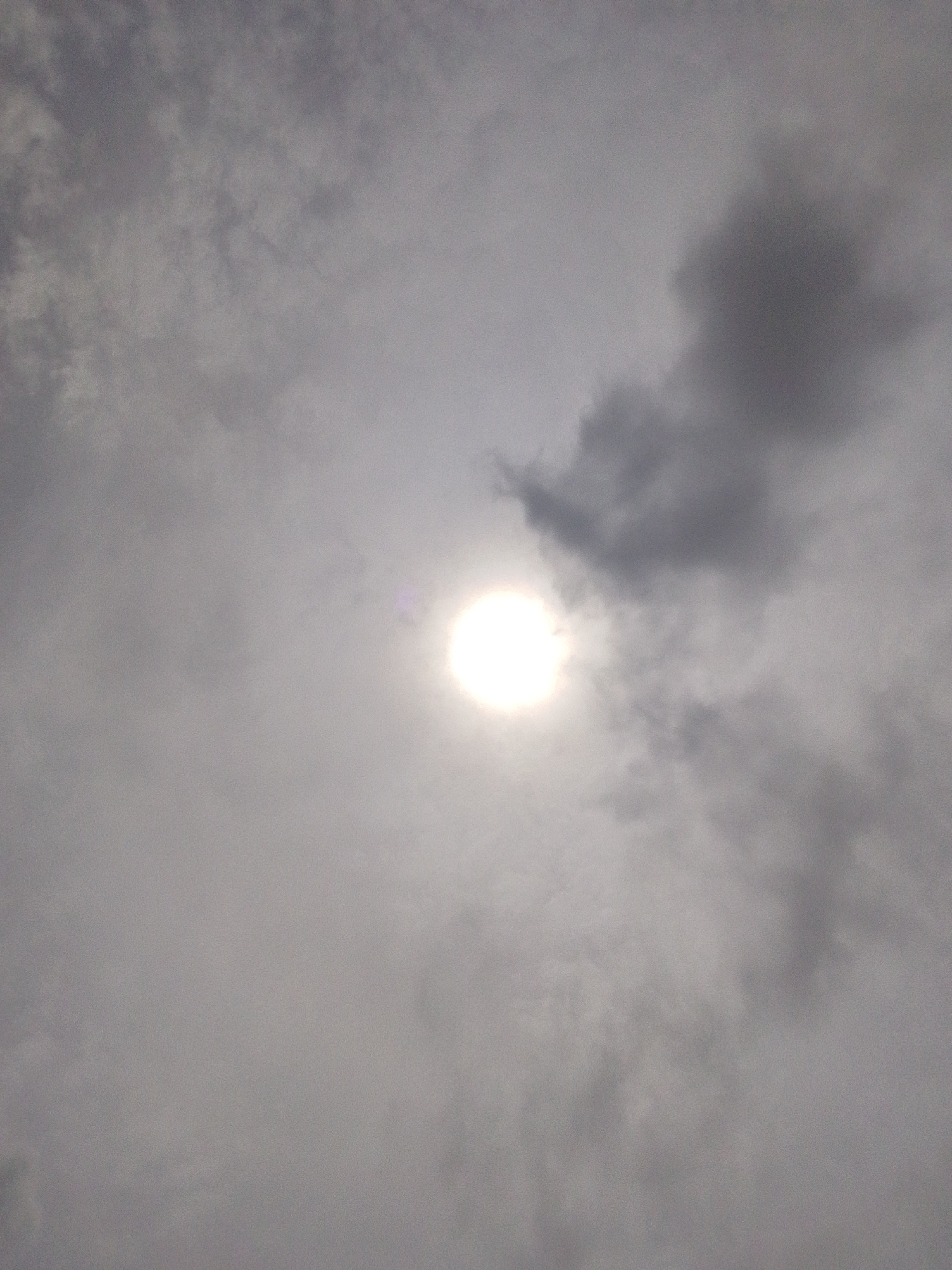 The image shows a cloudy sky with the sun partially visible as a bright, white orb. The clouds are a mix of light and dark gray, suggesting a partly cloudy or overcast day. There is a slight lens flare visible near the sun. The scene is devoid of any human presence or man-made structures. The environment is solely the sky. The location is specified as Serti, Nigeria.