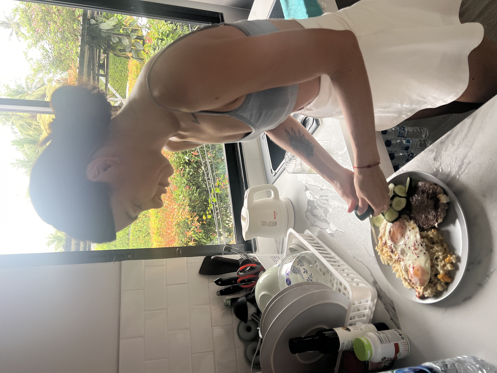 A woman in a grey sports bra and white skirt stands in a kitchen in บ้านบางรักษ์, Thailand, facing a plate of food. The plate contains two fried eggs, rice, steak, and sliced cucumbers. She is cutting a cucumber with a knife. A white kettle is on the countertop next to a dish rack filled with plates and bowls. Bottles of water are visible on the countertop and below the plates. Outside the window is lush greenery. The kitchen has white tile walls.