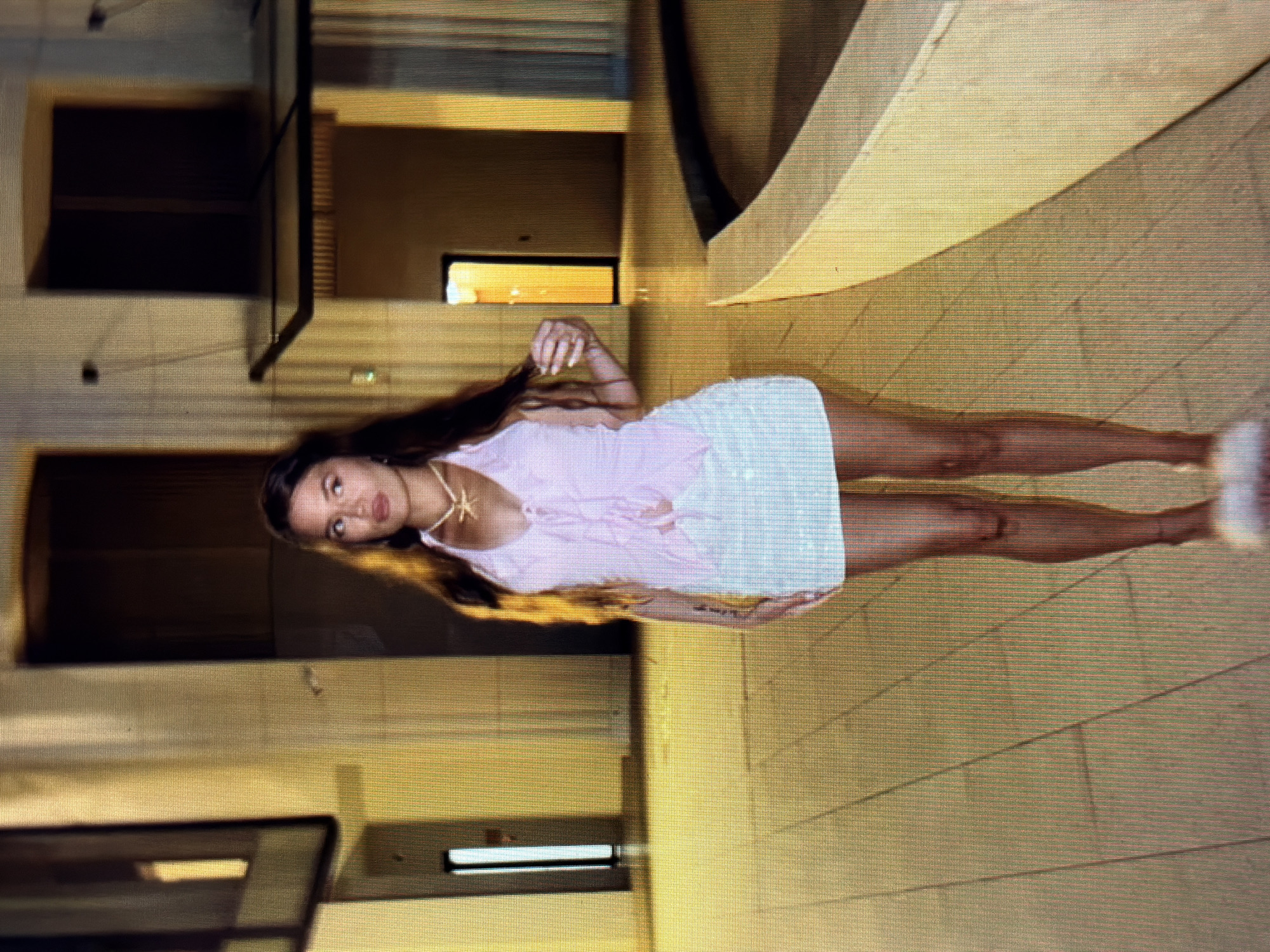 A woman stands in what appears to be an outdoor courtyard at night. She is wearing a light pink halter top with a tie in the front and a short, white, sequined skirt. She has long, dark hair and is wearing a light-colored necklace. The ground is paved with large, square tiles. Behind her, there are tall, cream-colored columns and dark doorways. To her right, there is a curved, light-colored structure, possibly a planter or fountain. The overall lighting suggests artificial illumination, casting long shadows. The image does not provide any information that would indicate a location in Sirius, Russia, or any specific city.