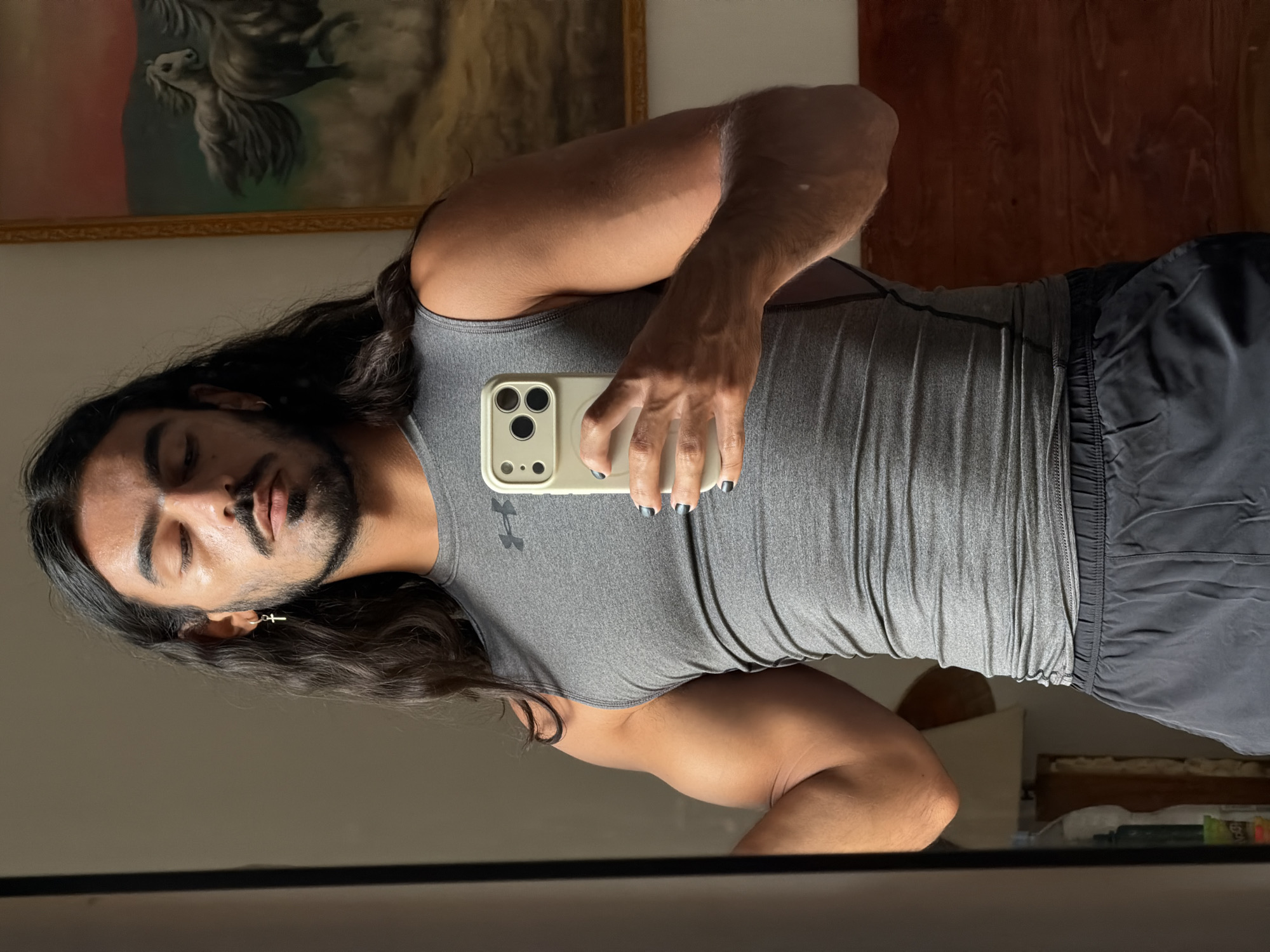 A man with long, dark hair and a beard is reflected in a mirror, holding a smartphone and posing. He is wearing a gray sleeveless athletic shirt and dark shorts. He has a cross earring in his left ear and dark nail polish on his fingernails. In the background, a painting of horses is visible. The setting appears to be a residential room in North Kuta, Indonesia.