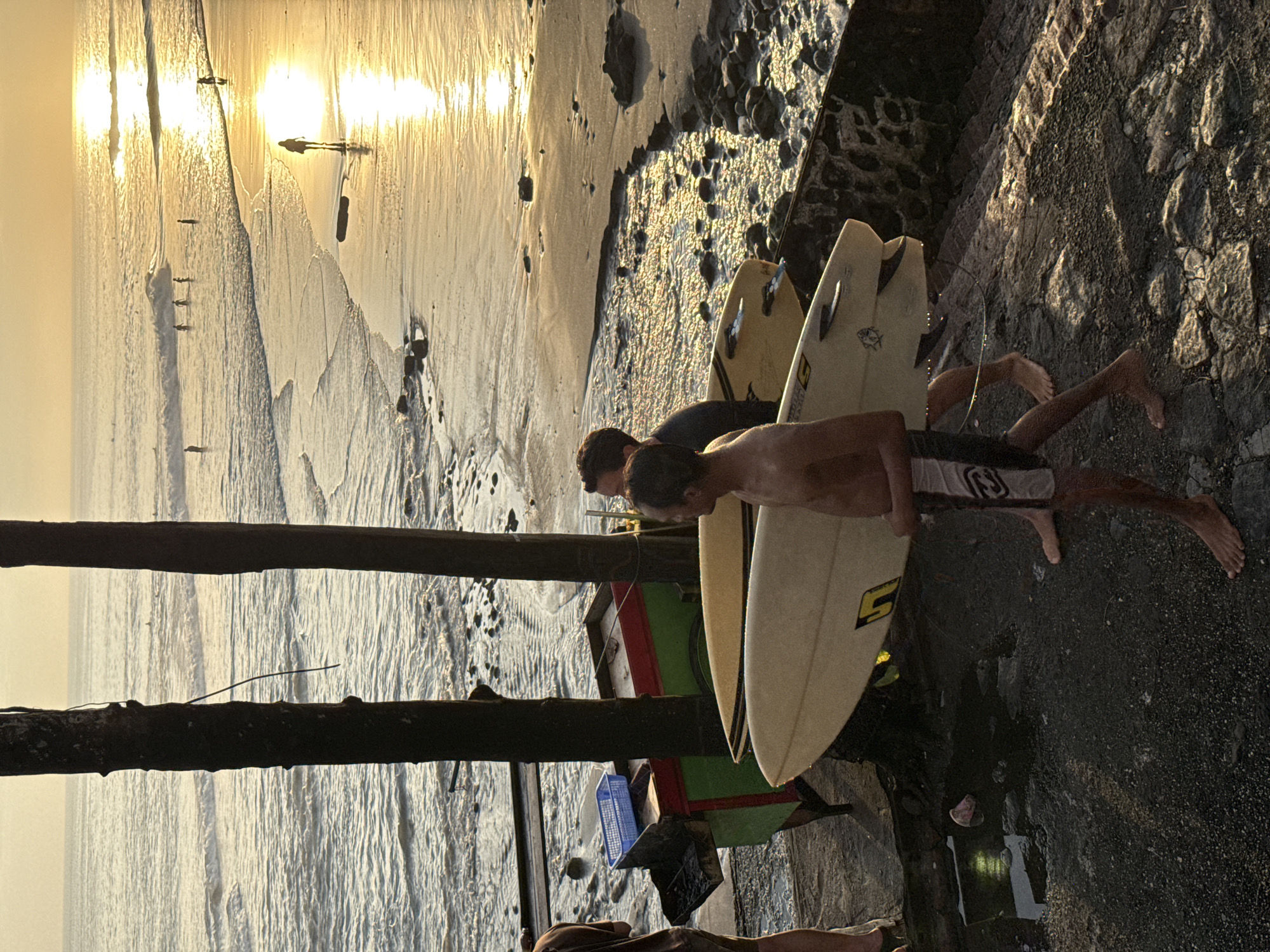 Two shirtless individuals are standing on a rocky surface, each holding a surfboard. The surfboards are leaning against a stone structure or embankment. Beyond the individuals and surfboards, there is a beach with sunlight reflecting off the wet sand. In the distance, there are small figures on the beach and in the water. The left side of the image has wooden posts. The scene is likely in North Kuta, Indonesia, as indicated by the beach landscape and possibly the style of the boats visible.