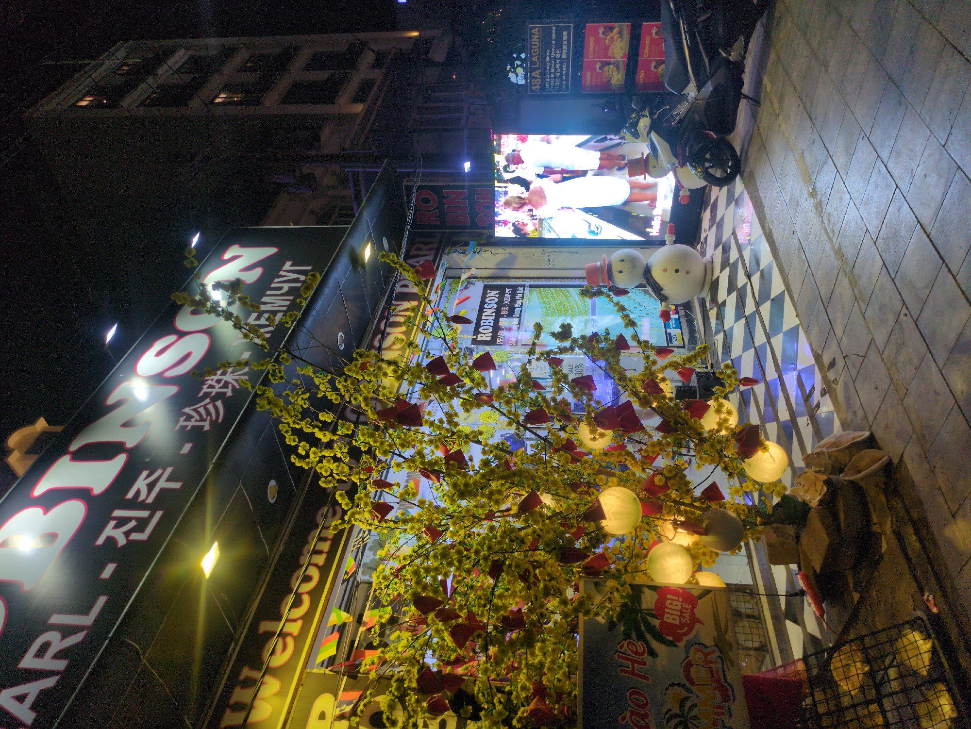 The image shows a street scene at night in Phú Quốc, Vietnam. A large sign with the text "BIRSEN PEARL - 진주 珍珠 EMЧУГ" is visible. Further down, there is a sign for "ROBINSON PEARL." Decorative trees with yellow flowers and red ornaments are in the foreground. A digital display screen shows an advertisement with people and text. A motorcycle is parked nearby on a black-and-white tiled sidewalk. The sidewalk leads to a grey-tiled area. Other signs in Vietnamese and English are present, including "48A LAGUNA" and "Chào Hè." Buildings are visible in the background.