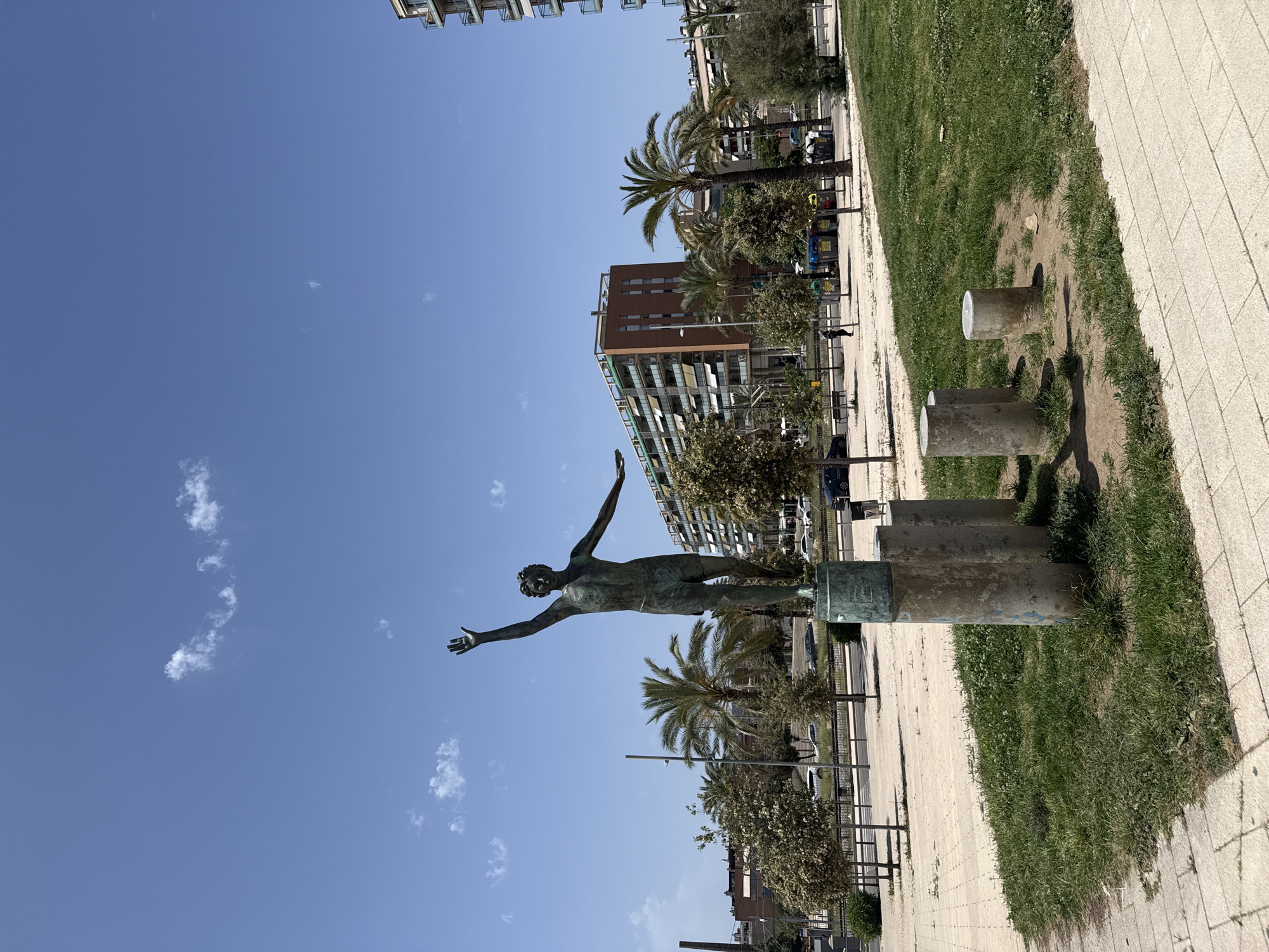 The image depicts a bronze statue of a male figure in athletic shorts, with his right arm raised and hand open, and his left arm extended outwards, posed as if in mid-stride or gesture. The statue is mounted on a cylindrical pedestal, which is surrounded by several other concrete cylinders of varying heights. This entire arrangement is situated on a patch of grass, bordered by a paved walkway and a street.
In the background, a clear blue sky with sparse white clouds stretches above a cityscape featuring several modern buildings. One prominent building is a tall, reddish-brown structure with multiple balconies and awnings. To its right, a taller, white building with numerous balconies is visible. Palm trees and other trees are scattered throughout the scene, some framing the buildings and others lining the street. Cars are parked or driving on the street, and a few individuals are walking in the distance, including one person near a row of colorful recycling bins. The lighting suggests it is daytime, likely with bright sunlight. The location is identified as the Estatua del Nadador (Swimmer Statue) in Badalona, Spain, near the beach promenade.