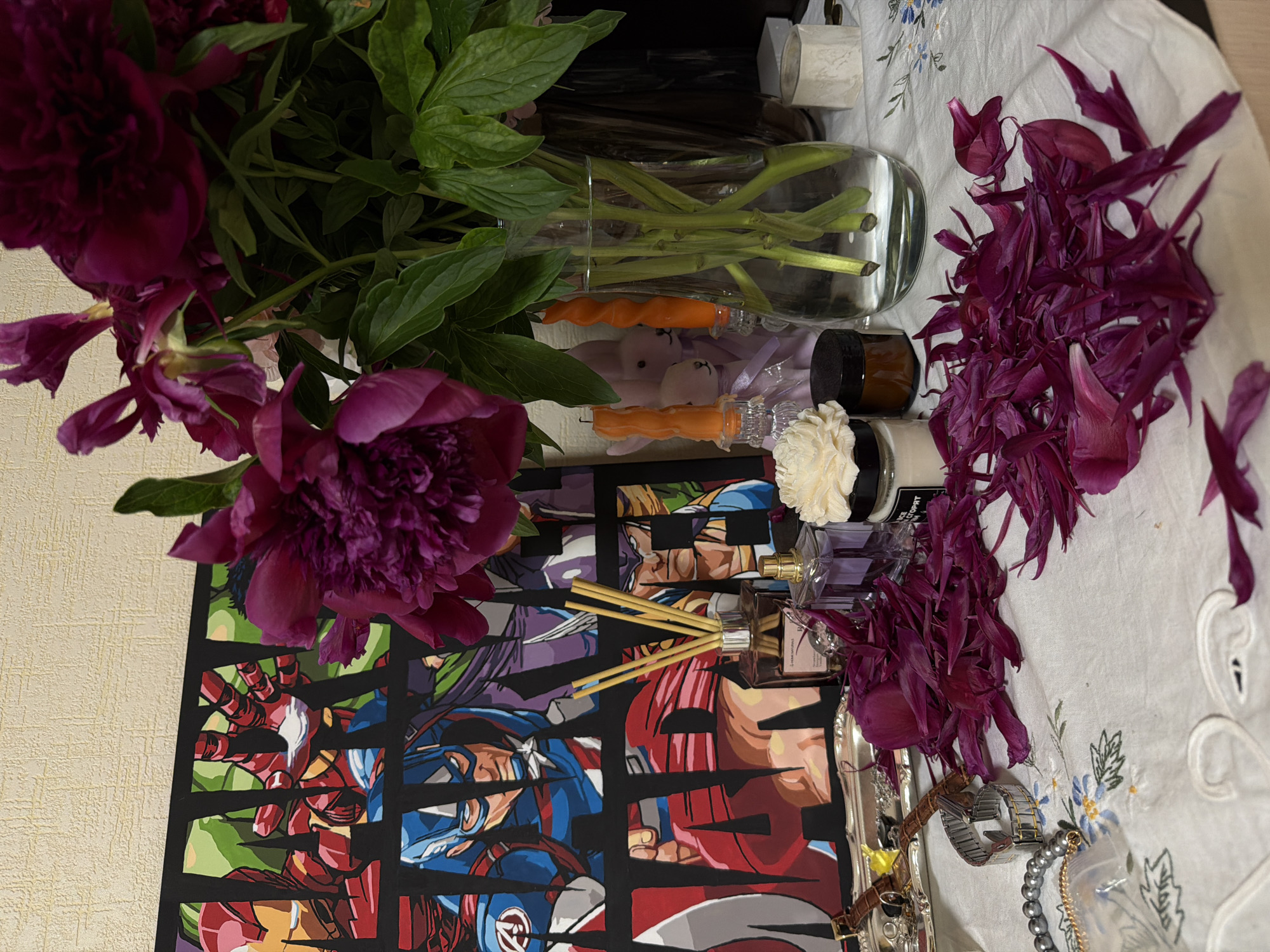 The image displays a flat lay composition of various objects arranged on a white tablecloth with delicate blue and green embroidered floral patterns. A large bouquet of dark magenta peonies with green leaves stands in a clear glass vase on the left. Numerous fallen dark magenta peony petals are scattered on the tablecloth to the right of the vase.
Below the vase, a black rectangular mat features colorful, stylized illustrations of fragmented popular culture characters, including Iron Man, Captain America, a Dragon Ball Z character (likely Goku), and Wolverine.
Other items on the tablecloth include two orange spiral candles in small glass holders, two small light pink or purple rabbit figurines, a small brown jar and a small white jar (both with black lids), and a white decorative flower. A clear glass diffuser bottle with reed sticks, labeled "L'ARTISAN PARFUMEUR Paris," is visible next to a clear glass perfume bottle with a golden cap. To the far right, a silver tray holds jewelry such as a silver watch, a grey beaded bracelet, and a pearl necklace.
The scene is an indoor setting, likely a table or dresser, showcasing a personal and aesthetic arrangement of items. No specific activity or event is taking place, and no time of day or weather is discernible.