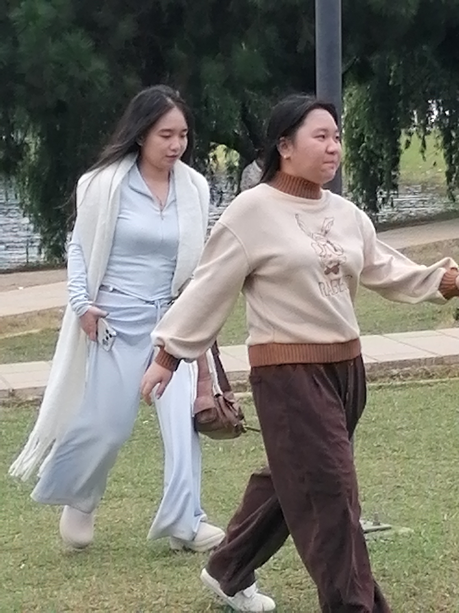 Two young women are walking on a grassy area with trees in the background. The woman on the left is wearing a long, light-colored scarf over a light blue hooded jumpsuit. She is holding a cell phone in her right hand. The woman on the right is wearing a cream-colored sweatshirt with a graphic of a rabbit and the word "RAGE" on it, and brown pants. She has a brown bag slung over her shoulder. The scene appears to be a park or outdoor recreational area, possibly in Da Lat, Vietnam, given the lush greenery and mature trees often found in the city's many parks and gardens. The women are walking away from the camera, with their bodies angled to the right.