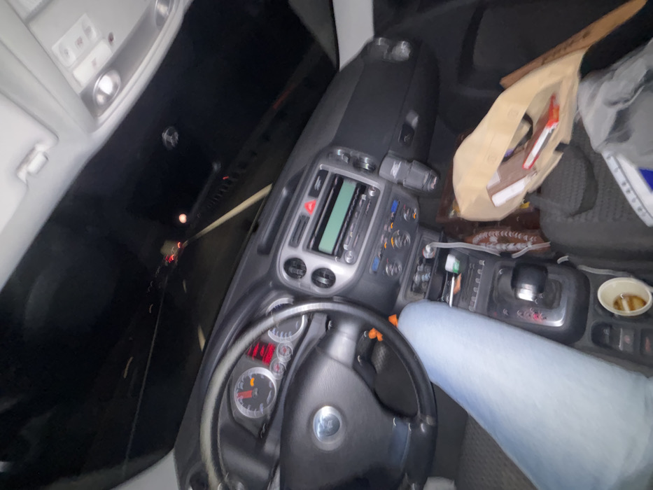The image depicts the interior of a vehicle at night in Pișchia, Romania, viewed from the passenger perspective looking forward. A driver's leg, clad in light blue jeans, is visible to the left of the center console. The black multi-function steering wheel with a Volkswagen emblem is positioned on the left. The instrument cluster behind the steering wheel displays illuminated red and orange indicators. The center dashboard features an infotainment system with a green-lit display, flanked by air vents and climate controls; the hazard light button is illuminated. Below, an automatic transmission shifter is visible, alongside various control buttons. A white cup containing a brown liquid rests in a cup holder in the center console. The front passenger seat is partially obscured by a tan paper bag containing miscellaneous items and a grey fabric. Through the windshield, a dark, unlit external environment is visible, with a road marked by a prominent white line extending into the distance. Multiple red taillights from other vehicles are visible in the far distance, suggesting traffic on a highway or main road, along with a blurred light-colored barrier on the right side of the road. The vehicle's interior ceiling is light-colored, showing overhead lights and controls.