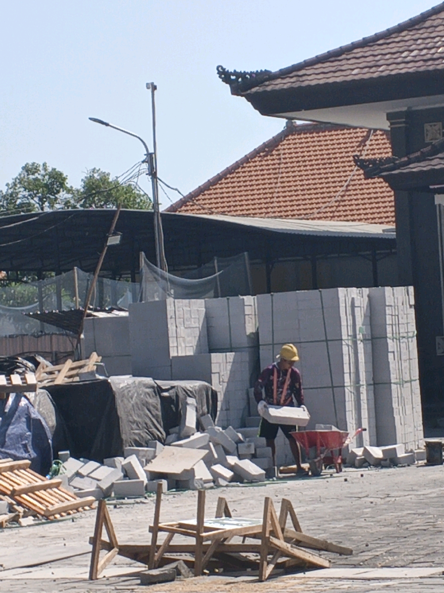 The image depicts a construction site in Denpasar, Indonesia, with stacks of large, light-colored concrete blocks, some wrapped in green strapping, dominating the mid-ground. In the foreground, a construction worker wearing a yellow hard hat, a maroon patterned long-sleeved shirt with orange suspenders, black shorts, and white gloves is carrying a single concrete block. The worker is standing next to a red wheelbarrow, with a pile of scattered concrete blocks and some wooden pallets nearby.
In the background, a building with a distinctive red tiled roof and ornate dark trim is visible. To the left of the building, a dark tarp covers some materials, and a light pole with a street lamp stands tall. Trees with green foliage can be seen behind the tarp. The ground is paved with a light-colored, textured surface, likely concrete or asphalt, typical of outdoor construction or industrial areas. The sky is clear and blue, suggesting fair weather. The overall scene is one of ongoing construction work, with materials neatly stacked and a worker actively engaged in moving a concrete block.