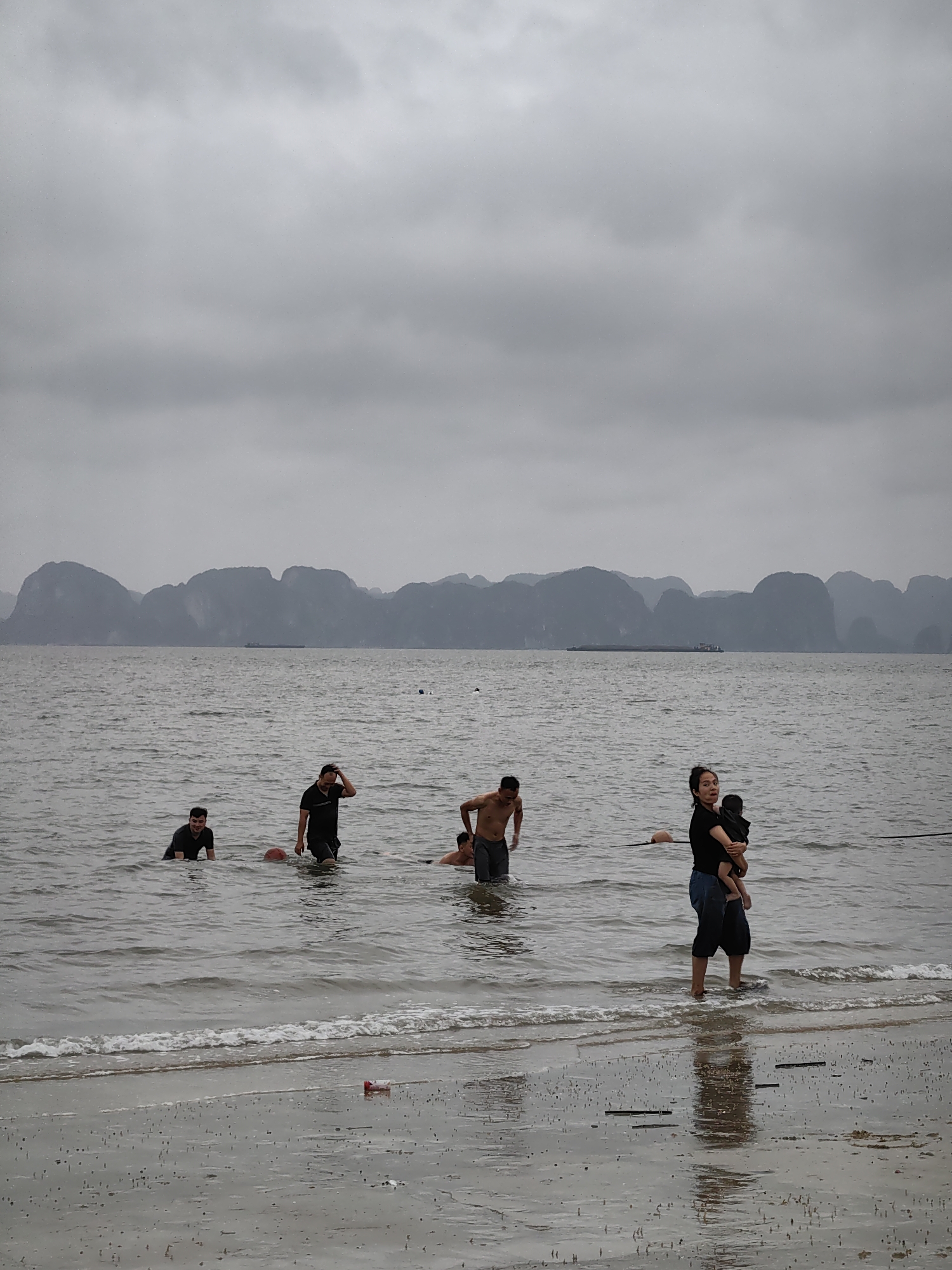 The image depicts a tranquil, overcast day at a beach or bay, characteristic of Bai Chay Ward, Vietnam, with distant limestone karsts dominating the horizon. The sky is a uniform, light gray, indicating heavy cloud cover.
In the foreground, a wet, reflective sandy shore is visible, marked by small ripples and gentle waves breaking at the water's edge. A discarded red and white plastic container lies on the sand. On the right, a woman in a black shirt and dark pants wades in the shallow water, holding a baby wrapped in dark fabric. She looks towards the camera with a neutral expression. A dark rope or line stretches across the water in front of her.
Further out in the sea, several men are present. On the left, a man in a dark shirt and shorts is partially submerged in the water. Towards the center, another man, also in a dark shirt, wades near a red ball floating in the water. Behind him, another shirtless man is partially visible. The people appear to be either swimming or wading, enjoying the calm waters.
The mid-ground features the expansive, gray sea, which appears relatively calm with minimal choppiness. In the distant background, a dramatic natural landscape unfolds, with a continuous line of dark, irregular limestone karsts or islands rising from the water, creating a distinctive silhouette against the sky. A long, dark barge or ship can be faintly seen navigating the waters between these landforms. The overall scene conveys a serene, almost muted atmosphere due to the lack of direct sunlight. No visible text is present in the image.