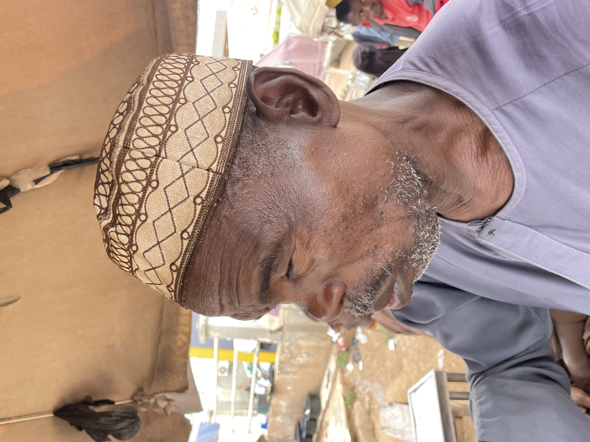 Here is a description of the image:
The image shows a close-up of an elderly man, facing slightly to the left, wearing a traditional patterned kufi cap. He is wearing a grey shirt and has a short, grey beard. The background suggests an urban environment, with hints of buildings and streets, which may be characteristic of Jos, Nigeria, based on the prompt. The focus is on the man's face, showing a weathered complexion and closed eyes.