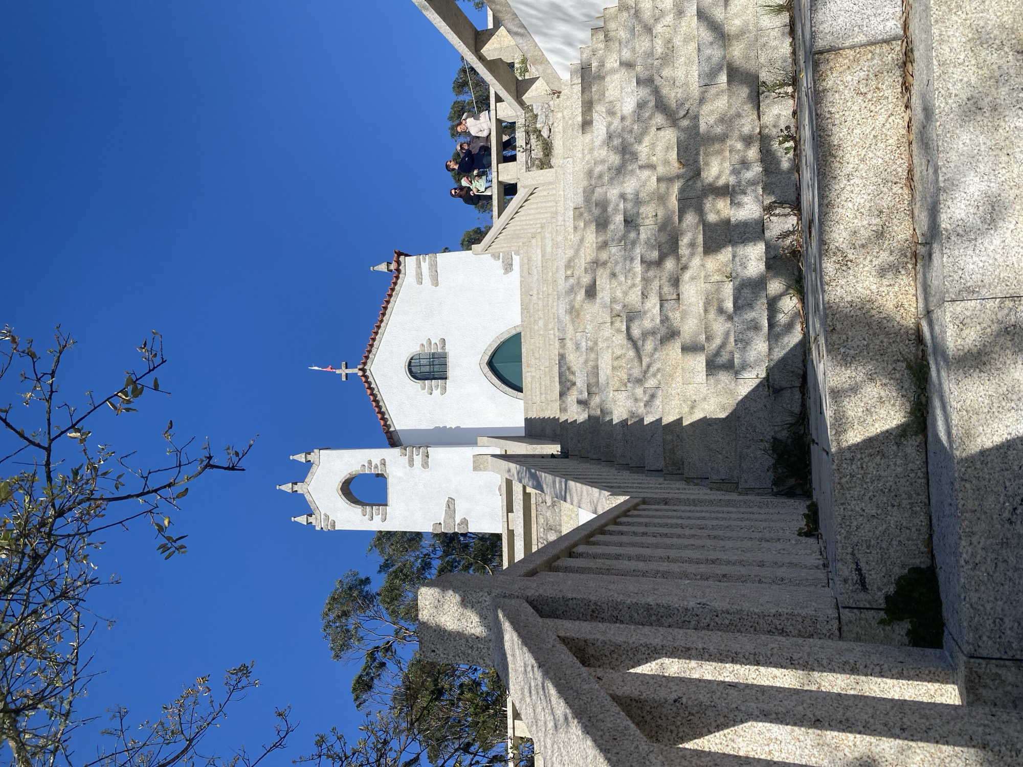 The image shows the Sanctuary of Monte da Saúde in Esposende, Portugal. A white chapel stands at the top of a long stone staircase. A group of people is visible at the top of the stairs near the chapel. The sky is clear and blue. Trees are visible in the foreground and behind the chapel. A stone railing lines the staircase.