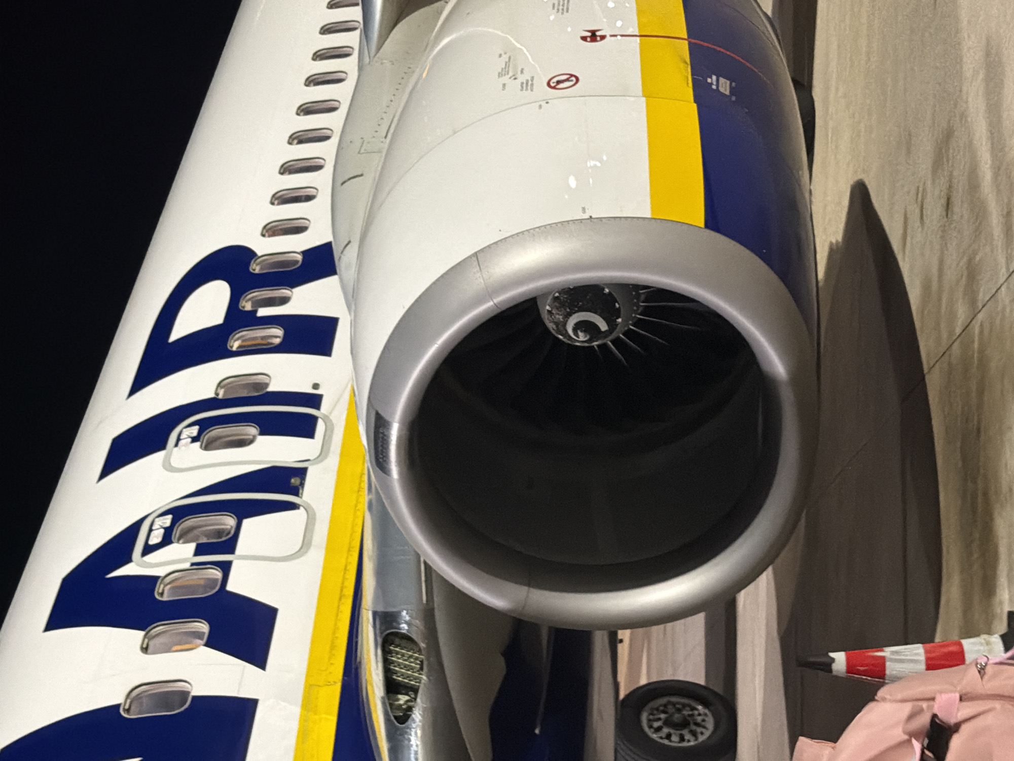 The image shows a close-up view of a Ryanair airplane's engine and fuselage at night. The aircraft is white with blue and yellow stripes. The letters "RYANAIR" are visible in blue on the fuselage, along with several windows. The large jet engine is prominently featured, with its fan blades visible inside. The scene appears to be on an airport tarmac. A small portion of a pink bag and a red and white striped cone are visible in the foreground, suggesting the presence of ground crew or passengers. The location is specified as Bari, Italy.