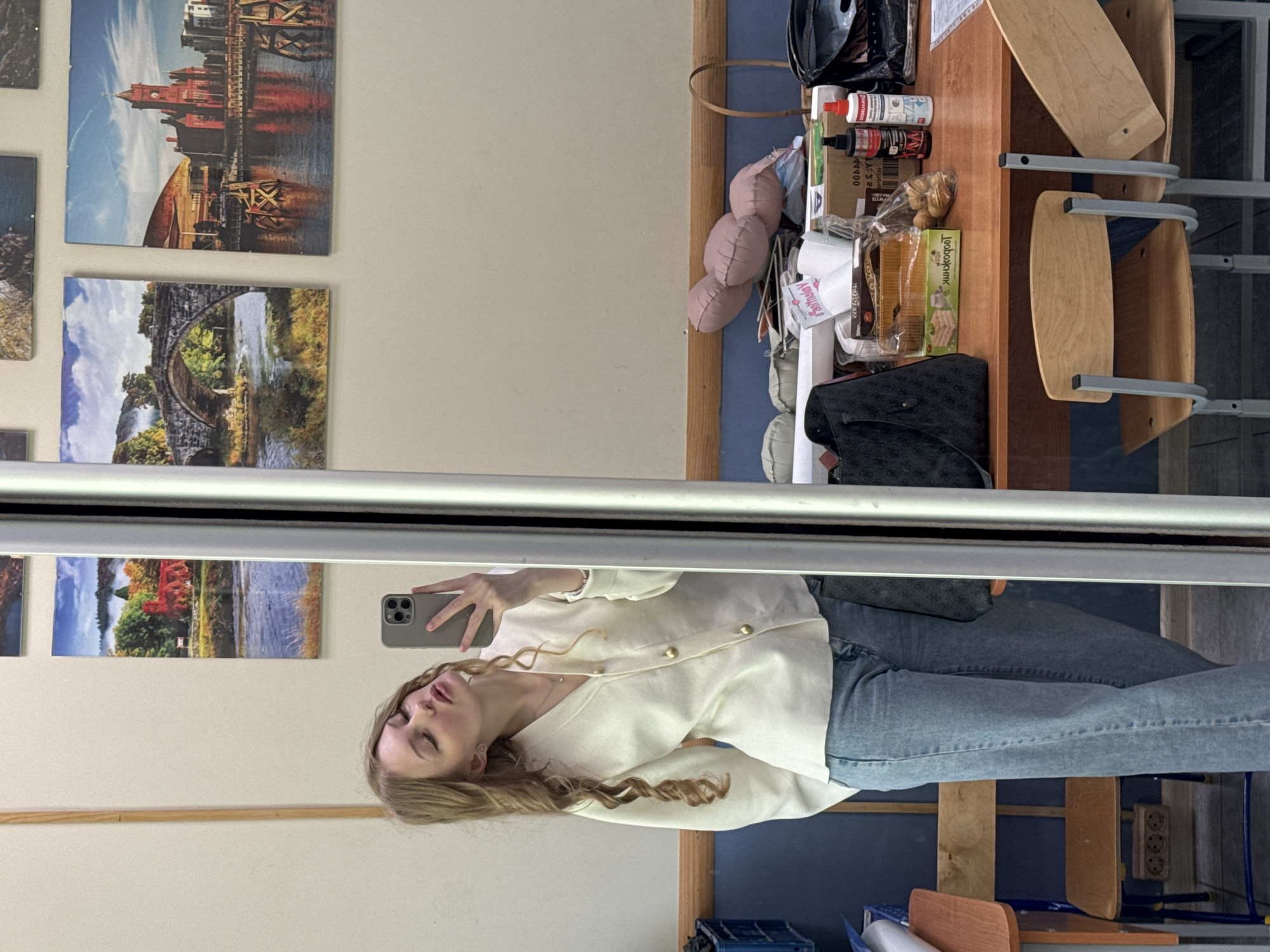 A mirror selfie depicts a person with long, wavy blonde hair wearing a cream-colored button-up shirt and blue jeans. The reflection shows a scene with artwork on the wall, a table with items including bottles, packaged food, and a black purse, and chairs stacked on top of each other. The location is not identifiable as Perm, Russia from the visual information provided.