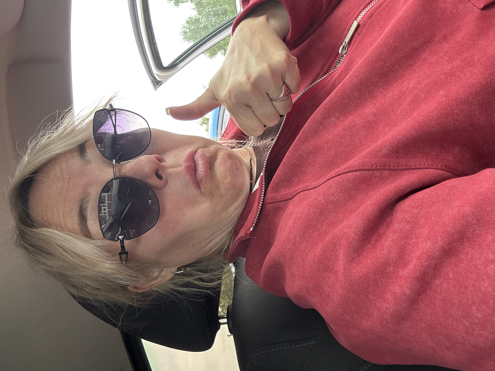 A woman is shown giving a thumbs-up in a car, with a slightly pursed expression and sunglasses on. She is wearing a red zippered hoodie and a ring on her thumb. The background shows the interior of the car and a slightly blurred view of trees through the open car door. The image appears to have been taken in daylight.