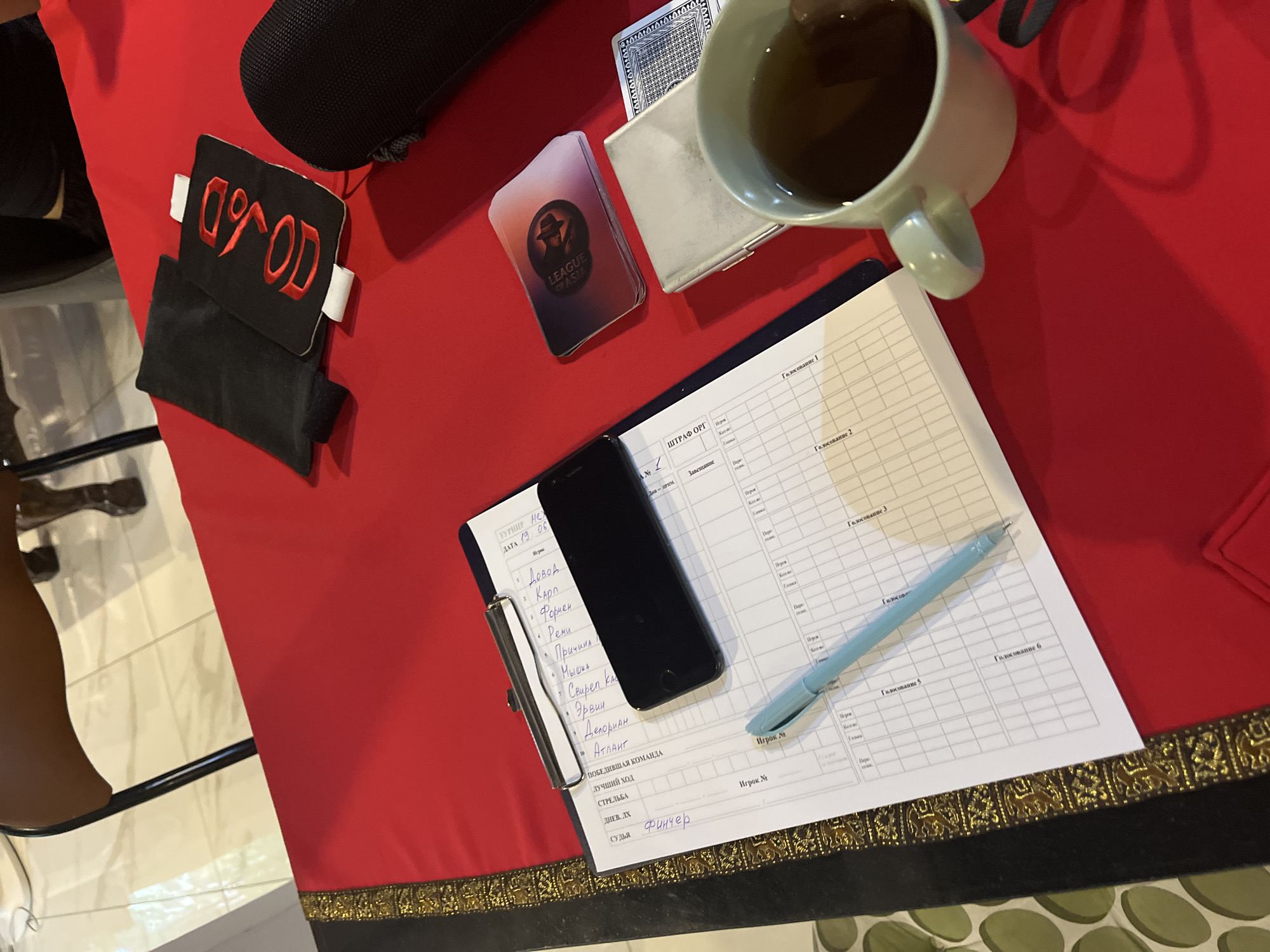 The image depicts a close-up view of a red tablecloth laid out on a table. The setting appears to be indoors, possibly a room in Chiang Mai City Municipality, Thailand, given the context provided.
On the table, several objects are arranged. In the foreground, a light green mug filled with dark liquid, likely tea or coffee, is visible. Next to the mug, a silver cigarette case and a deck of playing cards are placed. Further left, a clipboard holds several sheets of paper with handwritten notes and a list of names. A black smartphone rests on top of these papers. A light blue pen lies across the clipboard.
In the background, two black fabric pouches are laid out. One of these pouches has red embroidered lettering that reads "VOD". To the right of the pouches, a dark, textured cylindrical object, possibly a speaker or a case, is visible.
The lighting suggests an indoor environment, and shadows are cast on the red tablecloth. The overall impression is that of a table prepared for a game or a meeting, with items for scoring, communication, and leisure. The presence of a beverage and what appears to be a set of cards suggests a social gathering or a competitive event.