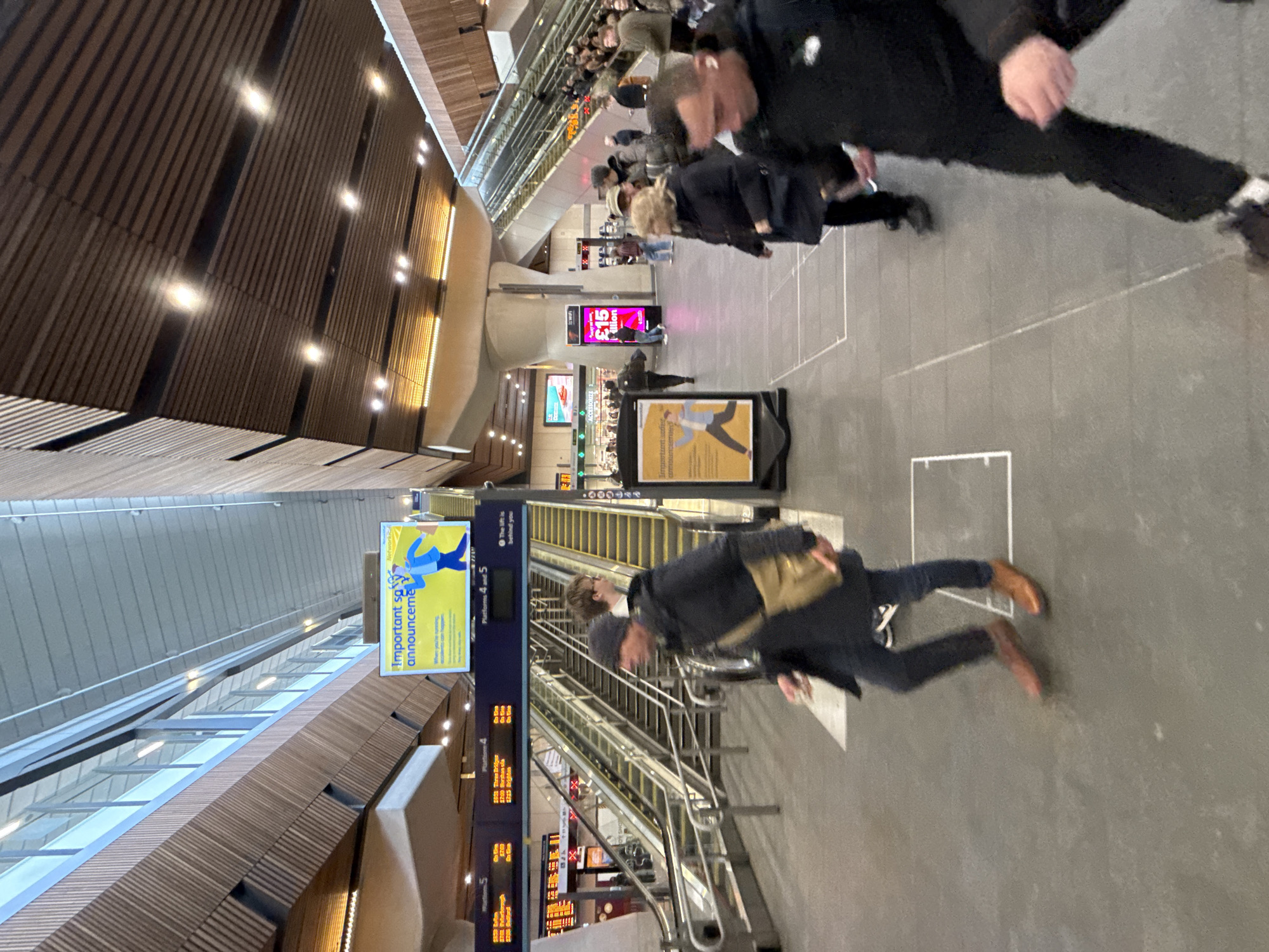 The photograph captures the interior of a transport hub, likely a train station, located in London, United Kingdom. The composition is taken at a slightly tilted angle. The image showcases a bustling scene with several individuals in motion, suggesting an active environment. In the foreground, several individuals are walking. Escalators are visible in the center of the frame. Signage is present, displaying information related to platforms, including platform numbers (4 and 5) and departure times to various destinations, such as "Three Bridges" and "Brighton". The walls and ceiling are wooden slats with embedded lighting. There are also advertisements and posters in the vicinity, with one particularly eye-catching poster advertising "£15" with the word "Million" below it. The overall ambiance suggests a contemporary transportation hub within a bustling urban setting.