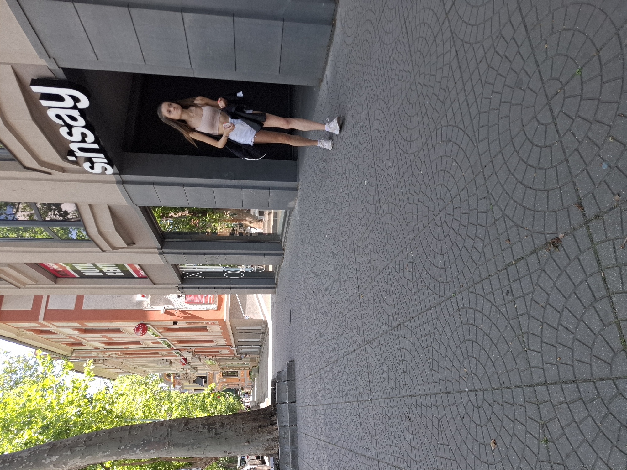 A young woman with long brown hair walks on a paved sidewalk in front of a Sinsay store. She is wearing a light pink crop top, white shorts, and white sneakers. She is carrying a black jacket over her left arm and a black bag over her shoulder. The Sinsay sign is prominent above the entrance. The sidewalk is paved with dark gray interlocking pavers arranged in a circular pattern. To the left of the sidewalk, there is a large tree trunk and a building with red and cream colored facades. The sky is bright and sunny, with green foliage visible from the trees.