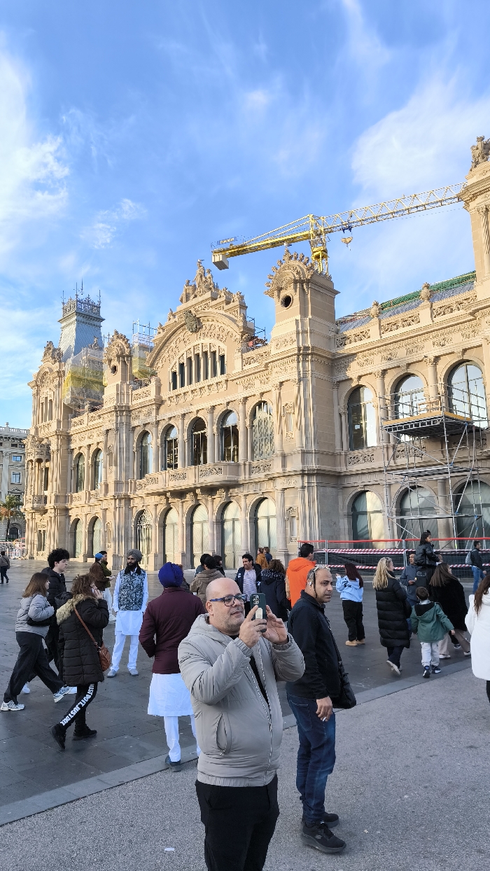 A man in a gray jacket takes a photo with his phone in front of a beige, ornately decorated building with scaffolding and a construction crane on top. Several other people walk around the plaza, including men wearing turbans and jackets, as well as women in winter coats. The sky above is blue with scattered clouds.