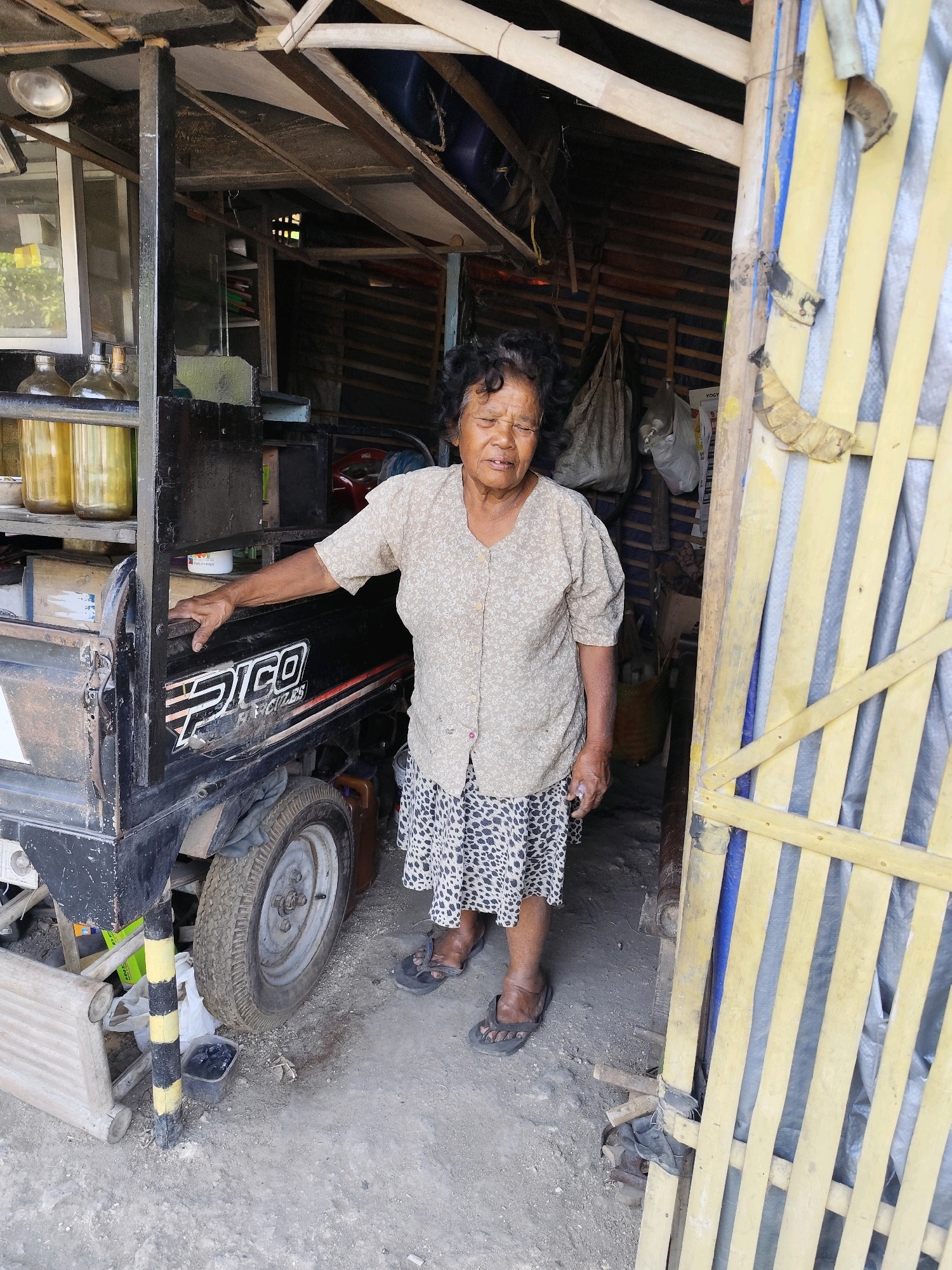 An elderly woman stands in the doorway of a makeshift structure in Bambang Lipuro, Indonesia. The structure appears to be made of wood and corrugated metal, with a yellow slatted gate on the right side. The woman is wearing a light-colored, patterned blouse and a leopard-print skirt. She is barefoot in dark-colored sandals. To her left is a black three-wheeled vehicle, possibly a modified motorcycle with a cargo bed. The vehicle has the word "PICO" written on its side. The ground is dirt and gravel. Behind the woman, inside the structure, there are various items visible, including bags, bottles, and shelves. The overall scene suggests a humble dwelling or small shop.