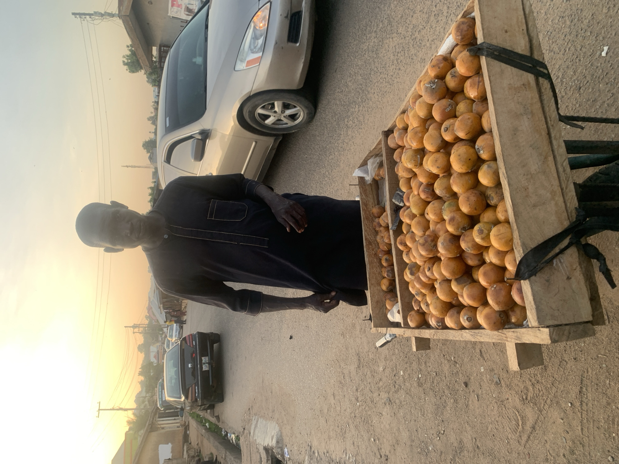 The image captures a street scene in Jos, Nigeria, featuring a man selling fruit from a cart during either sunrise or sunset. The sky displays warm, golden hues, indicating the soft light of dawn or dusk.
The primary subject is a man, positioned on the left side of the frame, dressed in a dark, long-sleeved tunic with subtle light stitching details. He is looking directly towards the camera with a neutral expression. To his right stands a wooden cart laden with numerous round, orange-yellow fruits, possibly a local variety of plum or African star apple, neatly arranged in wooden crates.
The setting is a street environment. A light-colored sedan, possibly a Honda Accord, is parked or moving slowly behind the fruit cart. Further down the street, a dark-colored sedan, likely a Peugeot, is also visible. The street surface appears to be a mix of asphalt and dirt/gravel. In the background, buildings and power lines extend along the road, contributing to the urban or semi-urban atmosphere.
The activity depicted is that of a street vendor, with the man standing by his fruit stall, ready to sell his produce. The overall impression is a typical roadside market scene in Nigeria. There is no clearly legible text on any signs or objects, though a partial license plate on the distant dark sedan shows "177" and some indistinct letters, consistent with Nigerian vehicle registration. The weather appears clear and calm.