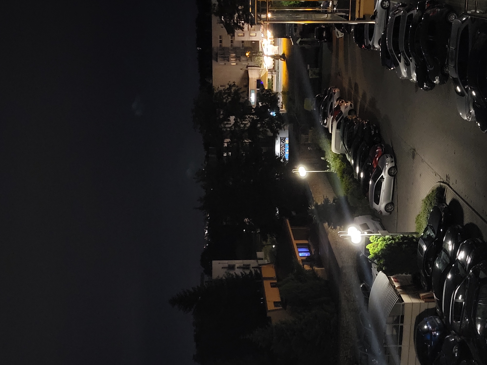 An aerial nighttime view captures a dimly lit parking lot filled with parked vehicles in a residential area. The scene is dominated by a row of dark-colored cars, with a few lighter-colored vehicles interspersed. Streetlights cast a glow over the asphalt, illuminating the vehicles and the surrounding environment. In the background, buildings and trees are visible, with some distant lights suggesting the presence of a city. The sky is dark and overcast, with a faint hint of clouds.