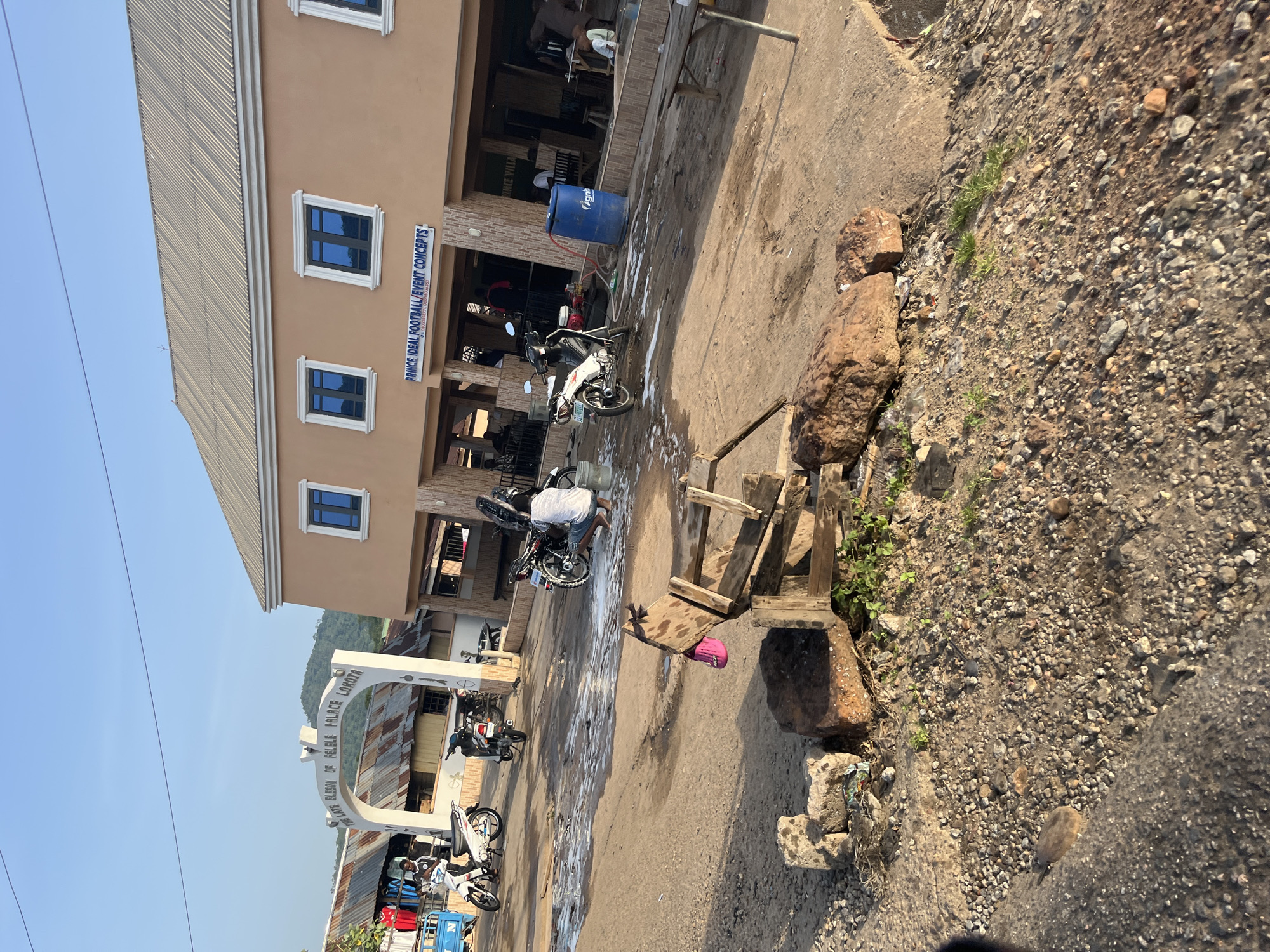 The image depicts an outdoor scene in front of a building with a tan facade and a corrugated metal roof. An arched gateway with text that reads "THE LATE EBASON OF FELELE PALACE LOKOTA" is visible on the left side of the image. Several motorcycles are parked in the foreground, with one being washed with soap suds. A person is crouched down, attending to the motorcycle. Another person is standing near a motorcycle in the background. The ground is a mix of dirt and gravel, with some scattered rocks and debris. In the distance, there are more buildings with tin roofs and some vegetation. The sky is clear and blue. A blue barrel is present near the building. A wooden structure with a pink shoe attached is in the foreground.
