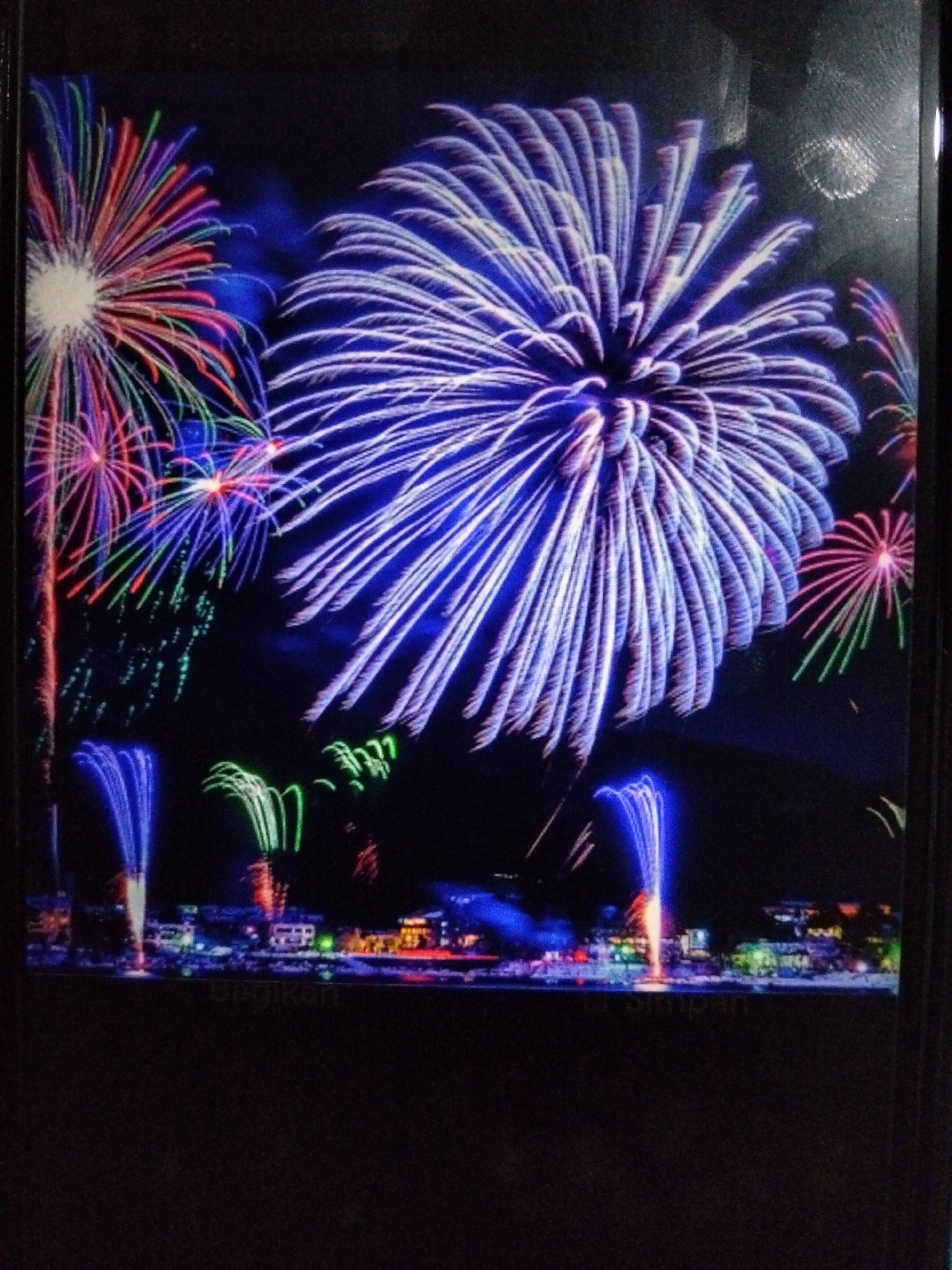 The image displays a vibrant fireworks display over a city at night. Large bursts of blue and white fireworks dominate the upper half of the frame, with streaks of red, green, and purple fireworks scattered throughout. In the lower portion of the image, a city skyline is visible, illuminated by the colorful explosions above. The city appears to be located in Kualaraja, Indonesia, as indicated by the watermark "Kualaraja, Indonesia" faintly visible in the foreground. There are no people or specific objects clearly discernible in the image besides the fireworks and the city. The overall scene evokes a sense of celebration and festivity.