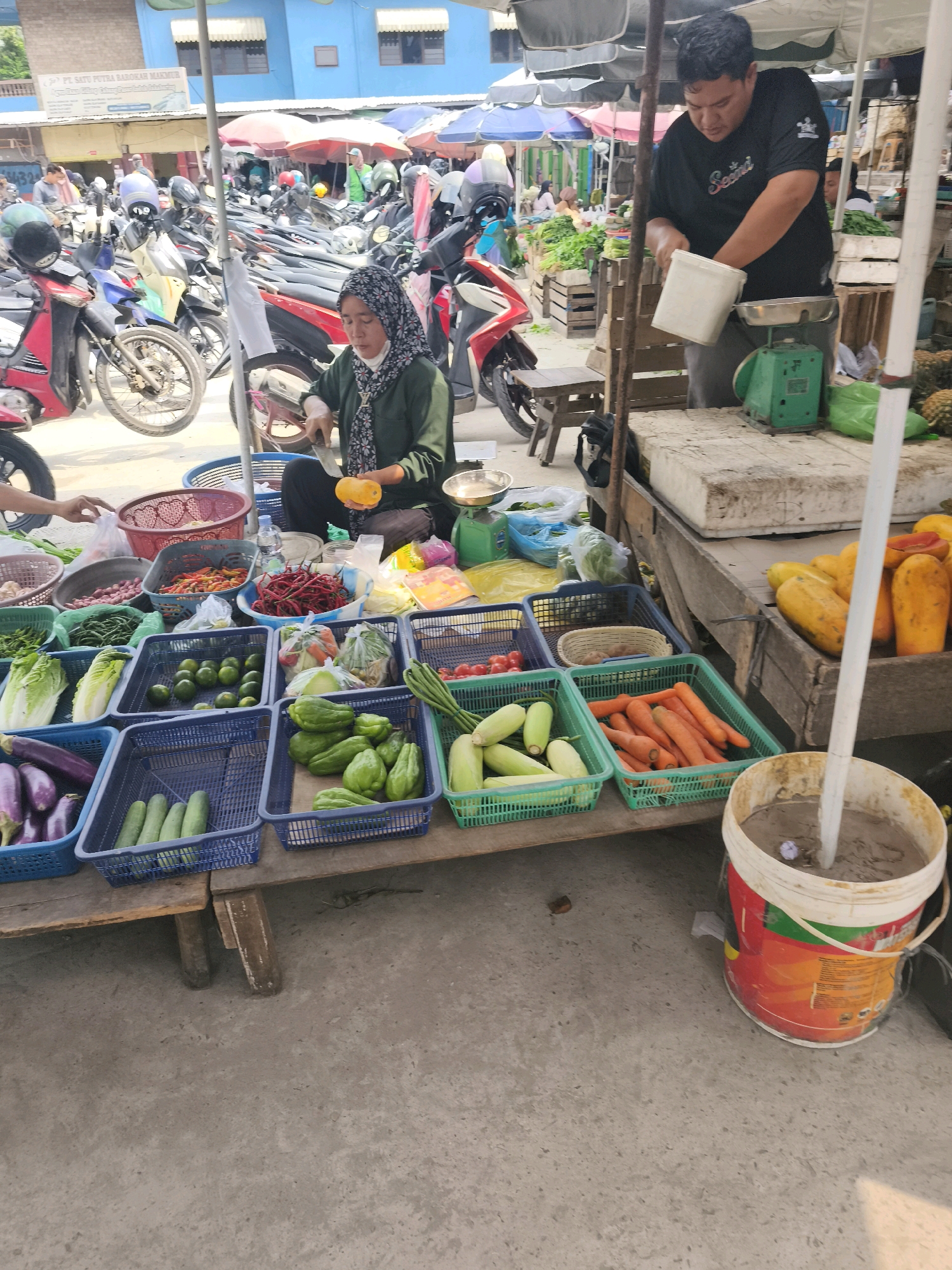 This image depicts a street market scene, likely in Palembang, Indonesia. In the foreground, a woman wearing a hijab and a green top is seated, preparing produce. She is holding a yellow fruit, possibly a papaya, and a knife, suggesting she is cutting or peeling it. Around her are various baskets and containers filled with fresh vegetables and fruits, including chili peppers, long beans, cucumbers, eggplants, limes, and carrots.
To the right, a man in a black t-shirt is standing and appears to be weighing produce on a green scale. He is holding a white bucket. Behind them, numerous motorcycles are parked, indicating a bustling commercial area. The market stalls are covered with awnings, providing shade from the sun. The ground is paved with concrete. In the background, buildings and more market activity are visible, hinting at the lively atmosphere of the location. The overall impression is one of a typical day at a vibrant Indonesian market.