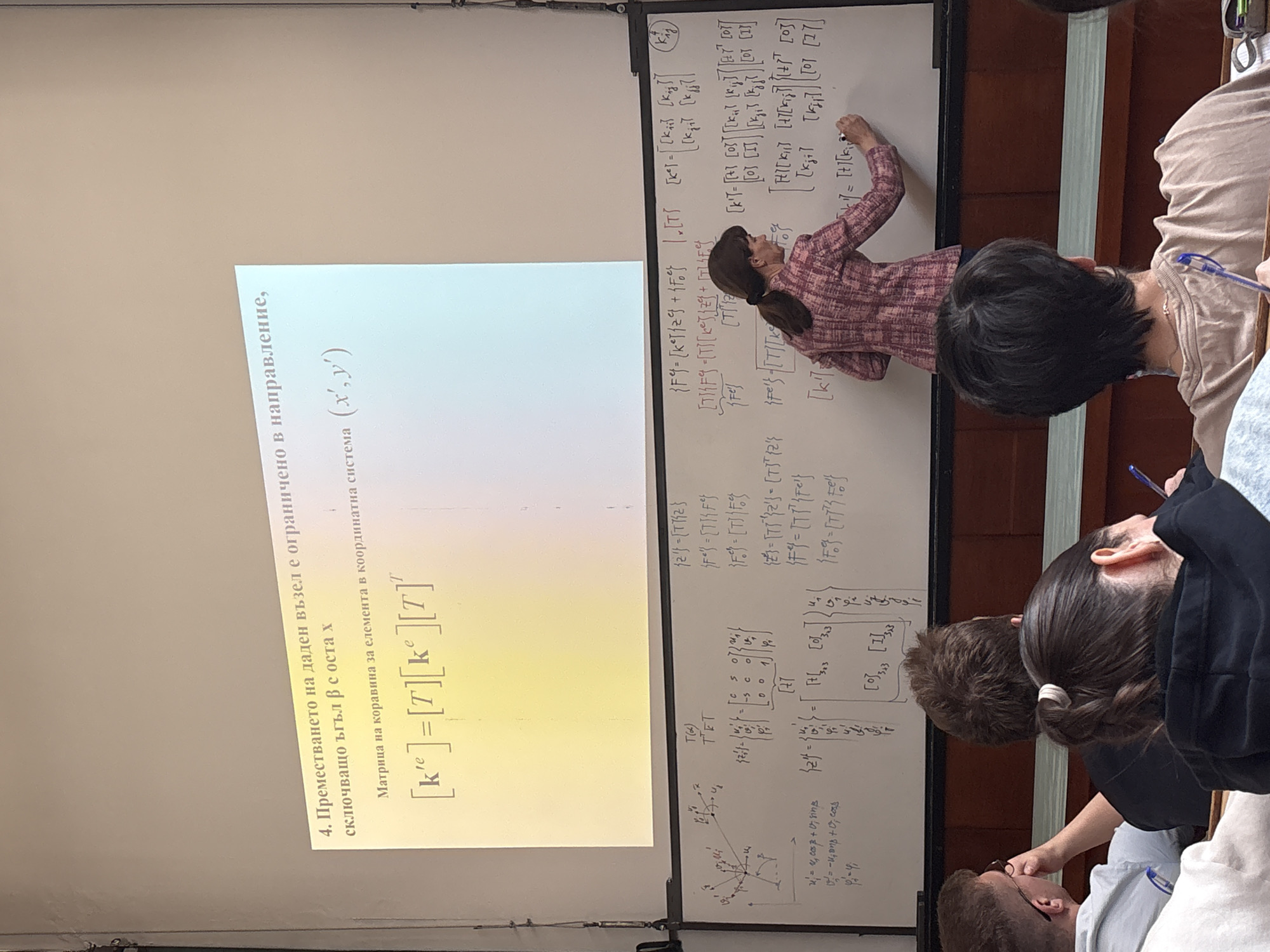 Here's a detailed description of the image:
**Scene:** The photo appears to be taken inside a lecture hall or classroom.
**People:**
* Several students are present, facing a whiteboard.
* A woman (likely the instructor or professor) is writing on the whiteboard. She is wearing a patterned top and has dark hair pulled back.
* One student is taking notes.
**Subjects:**
* Mathematical equations and explanations are written on the whiteboard.
* A presentation slide is being projected, with a title in Bulgarian related to displacement and stiffness matrices.
**Objects:**
* Whiteboard with equations and diagrams
* Projector screen displaying presentation slide in Bulgarian
* Writing utensil (pen or marker)
* Lecture hall environment with chairs/tables
**Actions:**
* The instructor is writing on the whiteboard, presumably explaining the concepts presented.
* Students are observing and potentially taking notes.
**Interactions:**
* The primary interaction is between the instructor and the students, with the instructor conveying mathematical information and the students receiving it.
**Environment:**
* The location appears to be an academic setting.
* The text from the presentation slide on the left side of the image is written in Bulgarian. According to the written text, the lecture is talking about "The displacement of a given node is limited in direction, including an angle β with the x axis. Stiffness matrix for the element in coordinate system (x',y')"
There is no definitive information in the image to pinpoint the exact location within Sofia. The presence of Bulgarian text suggests the photo was taken in Bulgaria. Given the academic context and mathematical content, it's highly probable that this is a lecture hall within a university or technical college in Sofia.