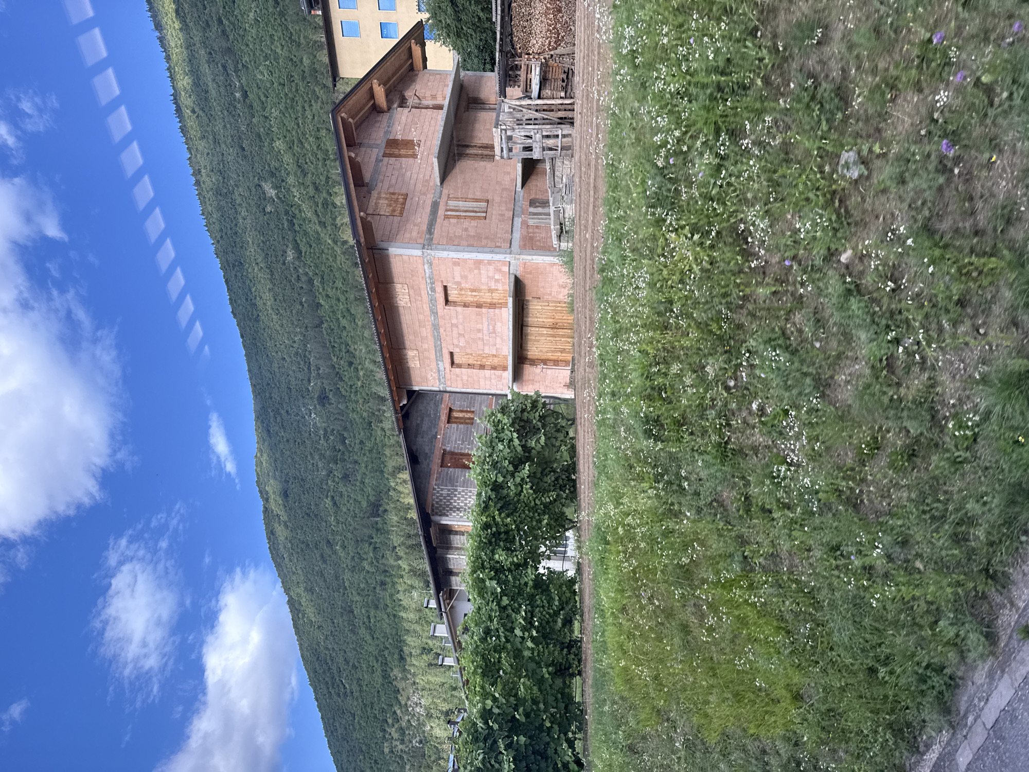 The image shows a view of buildings and a mountain landscape, likely from a moving vehicle given the streaking reflections on the blue sky. In the foreground, a grassy slope with scattered white and purple wildflowers descends towards a paved area. Behind the slope, a neat green hedge separates the vegetation from a construction site.
The construction site features two buildings. The one on the left is a long, low structure with a grey facade, seemingly unfinished, with wooden doors and window frames. To its right, a larger, multi-story building is under construction, built with pinkish-red bricks. This building has multiple wooden doors and boarded-up window openings. A scaffolding-like wooden structure and stacked lumber are visible near this building. Further to the right, a yellow building with blue windows is partially visible, suggesting it is a completed structure.
The background is dominated by a steep, tree-covered mountain that rises towards a bright blue sky dotted with white clouds. The reflection of square-shaped windows from the vehicle is visible in the sky. The overall scene suggests a rural or semi-rural setting in the Italian Alps. Without additional information, pinpointing the exact location within Spormaggiore is not possible from the image alone.