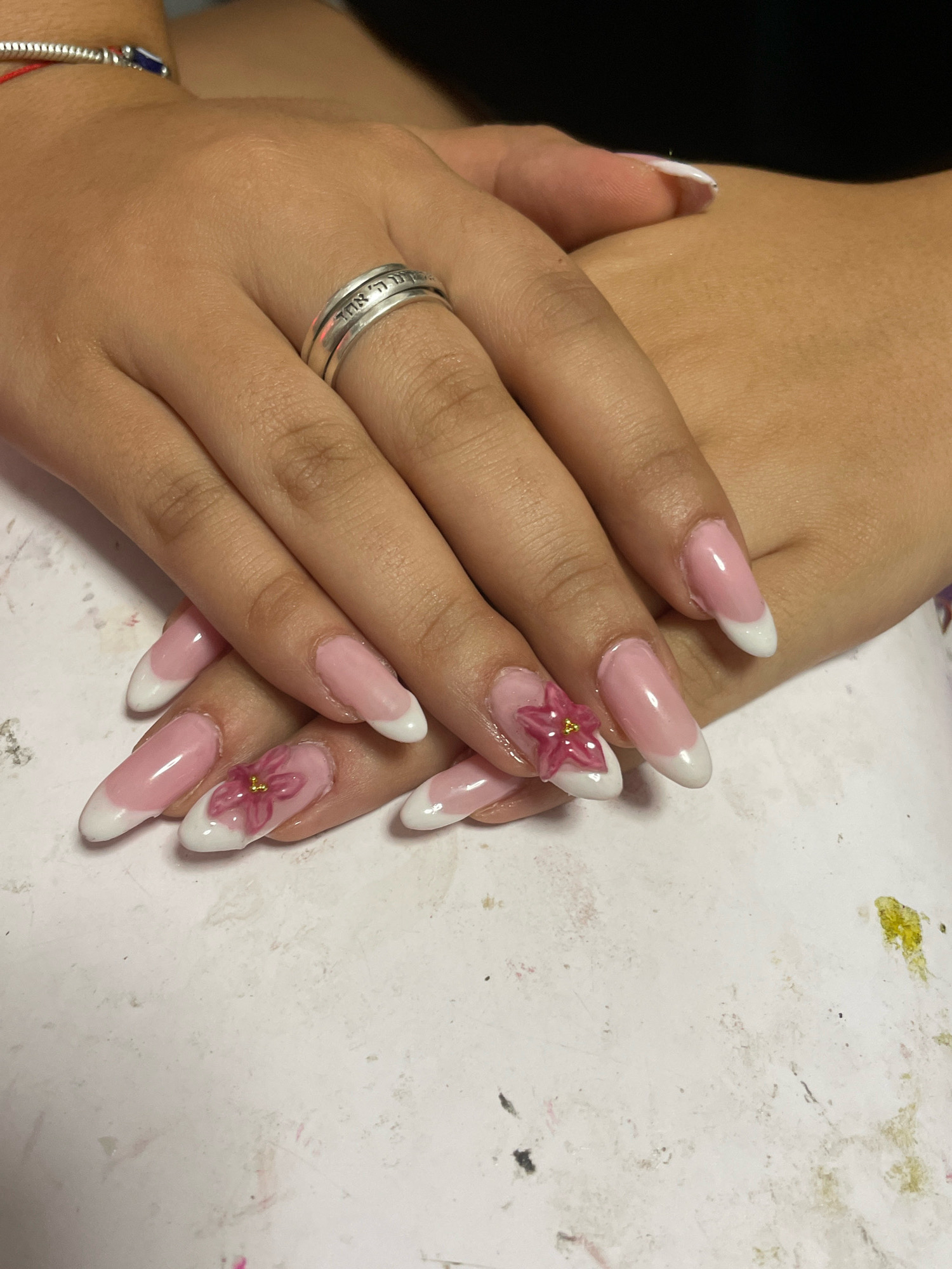 The image shows a close-up of a person's hands, displaying a manicure with pink and white French tips and a small, 3D pink flower with a gold center on one of the nails. The hands are resting on a white surface that appears to be a table or workspace, possibly in a nail salon. A silver ring with Hebrew inscriptions is worn on the ring finger. A silver charm bracelet with a red string and a blue charm is visible on the wrist of the left hand. The background is dark. The location is not specified in the image, therefore I cannot provide details about Raanana, Israel.