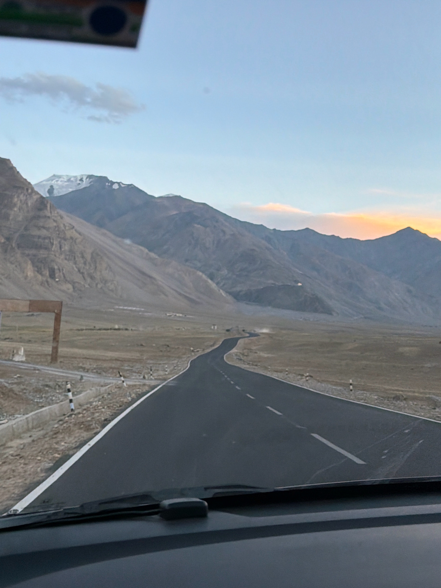 The image shows a winding asphalt road traveling through a desolate, mountainous landscape. The road is in the foreground, with the dashboard and windshield wipers of a vehicle visible at the bottom. The terrain is dry and barren, with sparse vegetation. In the distance, a range of large, rugged mountains forms the horizon. The sky is a pale blue with some wispy clouds, and the setting sun casts a warm glow on the mountain peaks. The overall impression is one of a remote and natural environment. The location is specified as Stongde, India, which is a remote region known for its monasteries and stunning mountain scenery. There are no people visible in the image.
