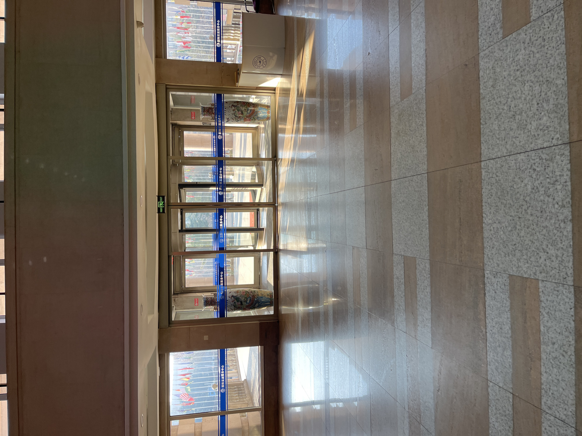 The image shows a hallway with a series of glass doors on the left side. The doors have blue vertical stripes and feature a logo that reads "国际交流服务中心" which translates to "International Exchange Service Center." Two porcelain vases with intricate designs can be seen through the glass doors. An "EXIT" sign is located above one of the doors. The hallway floor is covered with tiles in alternating light and dark shades. Light is streaming in from outside, creating a bright and reflective surface on the floor. The location is likely in Changchun City, China, based on the Chinese text.