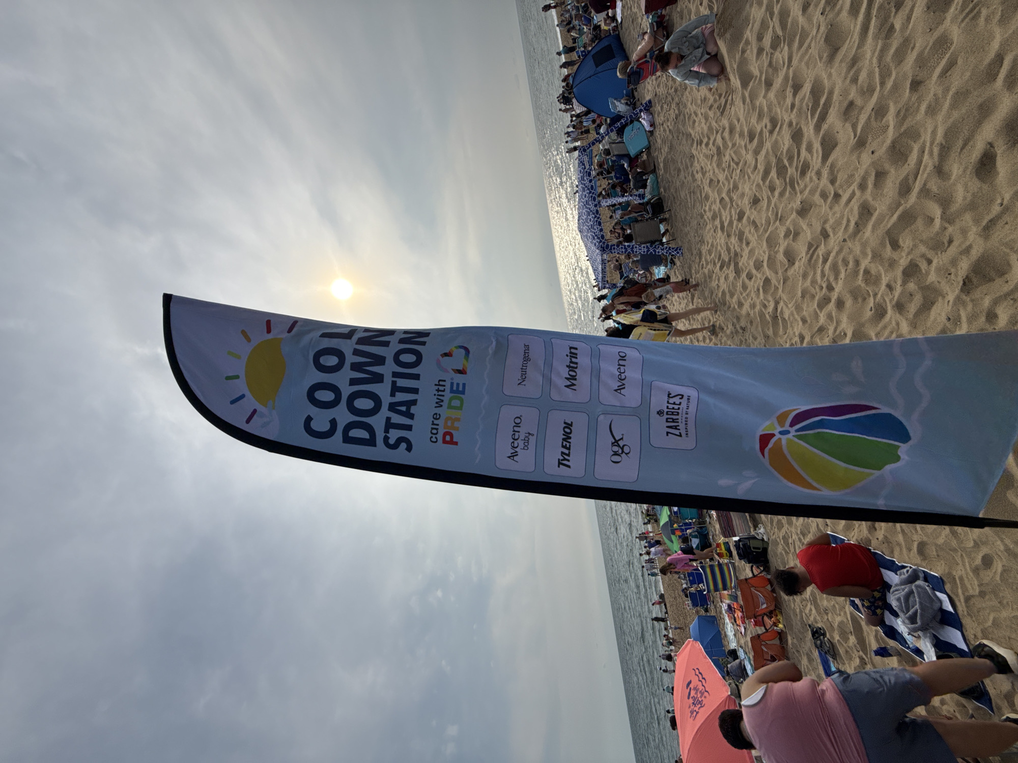 A light blue flag with the words "COOL DOWN STATION" printed in dark blue letters is positioned in the foreground of the image, partially obscuring the view of a crowded beach scene. The flag features a sun graphic and text that reads "care with PRIDE". Below this, several logos are displayed in a grid format, including Aveeno, Tylenol, OGX, Zarbee's, Neutrogena, and Motrin. At the bottom of the flag, a colorful beach ball graphic is visible.
The beach is populated with many people, some sitting in beach chairs, others lying on towels, and some walking around. There are various beach umbrellas and tents set up, providing shade. The ocean is visible in the background, with waves gently breaking on the shore. The sky is overcast with the sun shining through, creating a bright, hazy effect. The overall scene depicts a sunny day at a busy beach. Based on the context of a "Cool Down Station" with "PRIDE" branding and the presence of many people enjoying the beach, this scene is likely set in Provincetown, Massachusetts, known for its vibrant Pride celebrations and popular beaches.