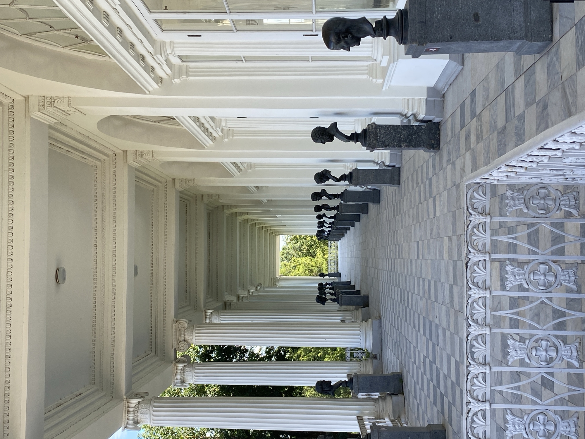 The image depicts the semi-circular colonnade of the Agate Rooms in Catherine Park, Pushkin, Russia. The colonnade features classical architecture with Corinthian columns supporting a white, ornate ceiling with decorative moldings. Along the tiled walkway, a series of dark stone pedestals display busts of historical figures. The busts are uniformly dark and appear to be made of bronze or a similar material, each rendered with detailed facial features. The walkway itself is paved with a checkerboard pattern of light and dark grey tiles. To the right, large arched windows are visible, reflecting the interior. The colonnade extends into the distance, creating a sense of depth and perspective. Trees and greenery are visible beyond the colonnade, suggesting an outdoor setting. A white decorative railing is in the foreground, partially obscuring the view of the walkway. The lighting is bright, indicating it is daytime. There are no people visible in the image.